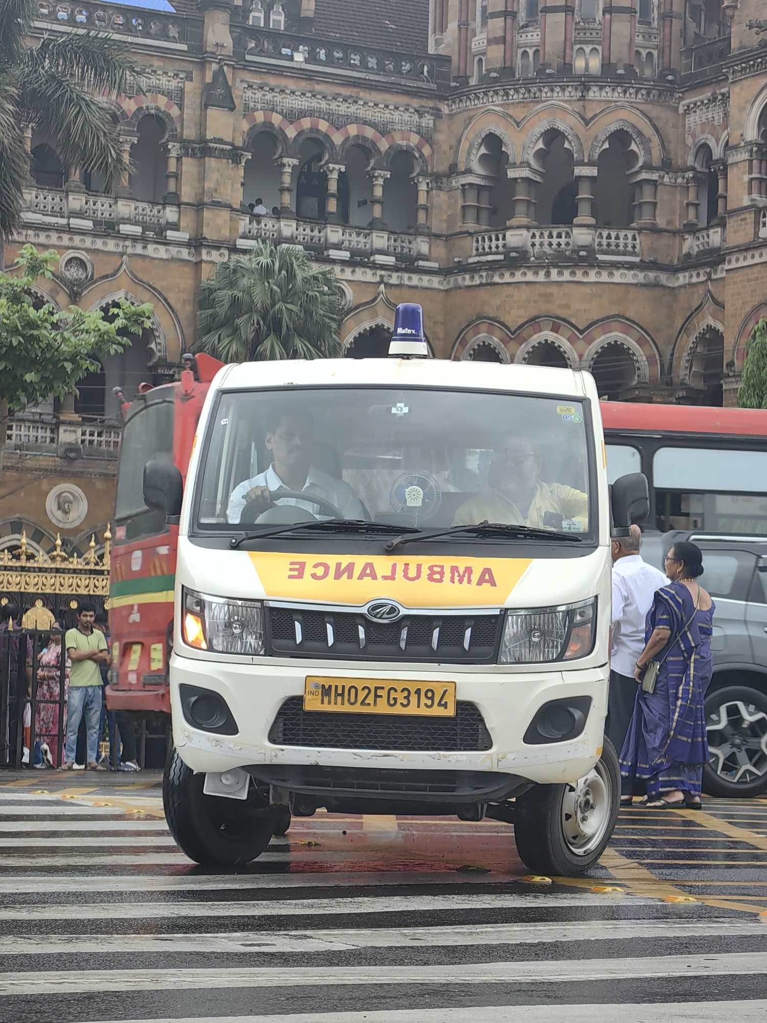 An ambulance is stopped at a pedestrian crossing in front of a large, ornate, historic building in Mumbai, India. The building features multiple arched windows, balconies with decorative railings, and a red-tiled roof. The ambulance is white with yellow and red lettering on the front that reads "AMBULANCE" in reverse. A blue flashing light is visible on its roof. The license plate reads "MH02FG3194".
Inside the ambulance, two men are visible. The driver, wearing a light blue shirt, is looking forward. A passenger in a yellow shirt is also in the front.
Several people are standing near the ambulance and on the sidewalk. A man in a green t-shirt and blue jeans stands on the left. A man and a woman in traditional Indian attire, a blue sari with gold trim, stand to the right of the ambulance. The woman is wearing a green crossbody bag.
Behind the ambulance, a red vehicle, possibly a fire truck or a bus, is partially visible. A dark grey SUV is parked to the right of the ambulance. The ground is wet, and the pedestrian crossing is marked with black and white stripes. Yellow raised markers are present on the crossing. Palm trees and other foliage are seen around the building. The overall lighting suggests an overcast day.