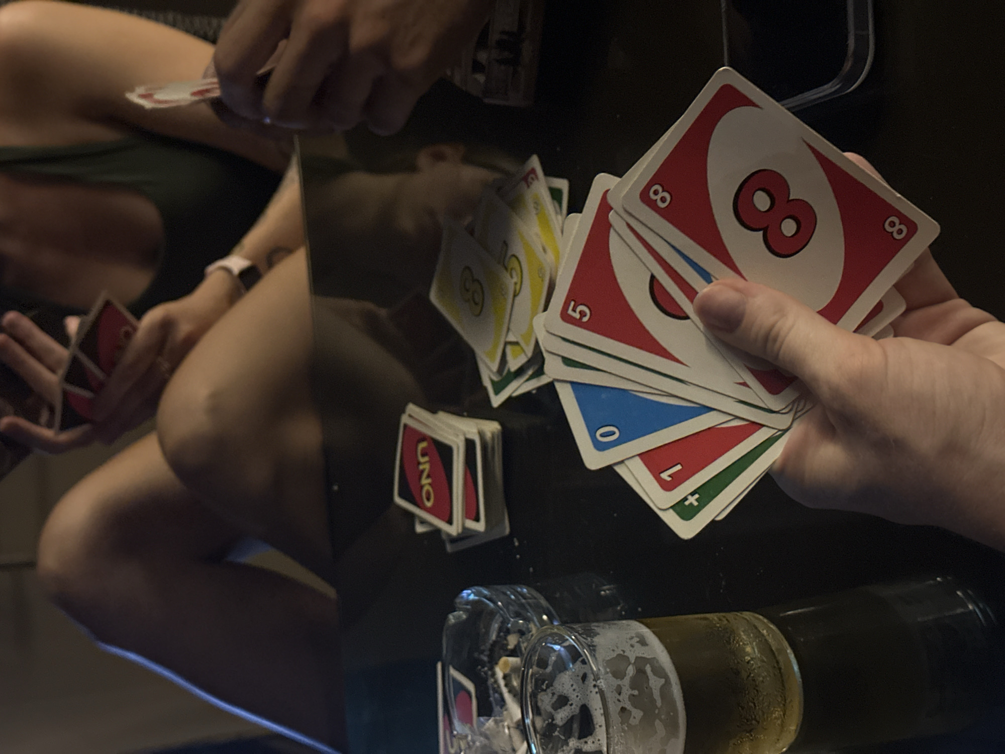 The image captures a casual, intimate scene of people playing Uno, likely in the evening or at night, in Baan Chaweng, Thailand. The primary focus is on the hands of two individuals and the Uno cards.
In the foreground, a hand holds a fan of Uno cards, prominently displaying a red '8'. Other cards in the hand show numbers '5', '0', and '1', in red, blue, and green, along with a green '+1' or '+2' card. On the dark, highly reflective table surface, a stack of Uno cards with the "UNO" logo on their backs is visible. Adjacent to the cards, a glass containing a foamy beverage, likely beer, is present, along with what appears to be another glass or an ashtray, and a beer bottle.
In the background, blurred reflections of cards and other hands are visible on the table. The torso and legs of another person can be seen, suggesting a close-up perspective of the game in progress. This person's hands are also holding cards. The lighting is dim, indicating an indoor setting during the evening hours. The reflective nature of the table creates a dynamic visual effect, mirroring the objects and people. The scene conveys a relaxed, social atmosphere centered around the card game and drinks.