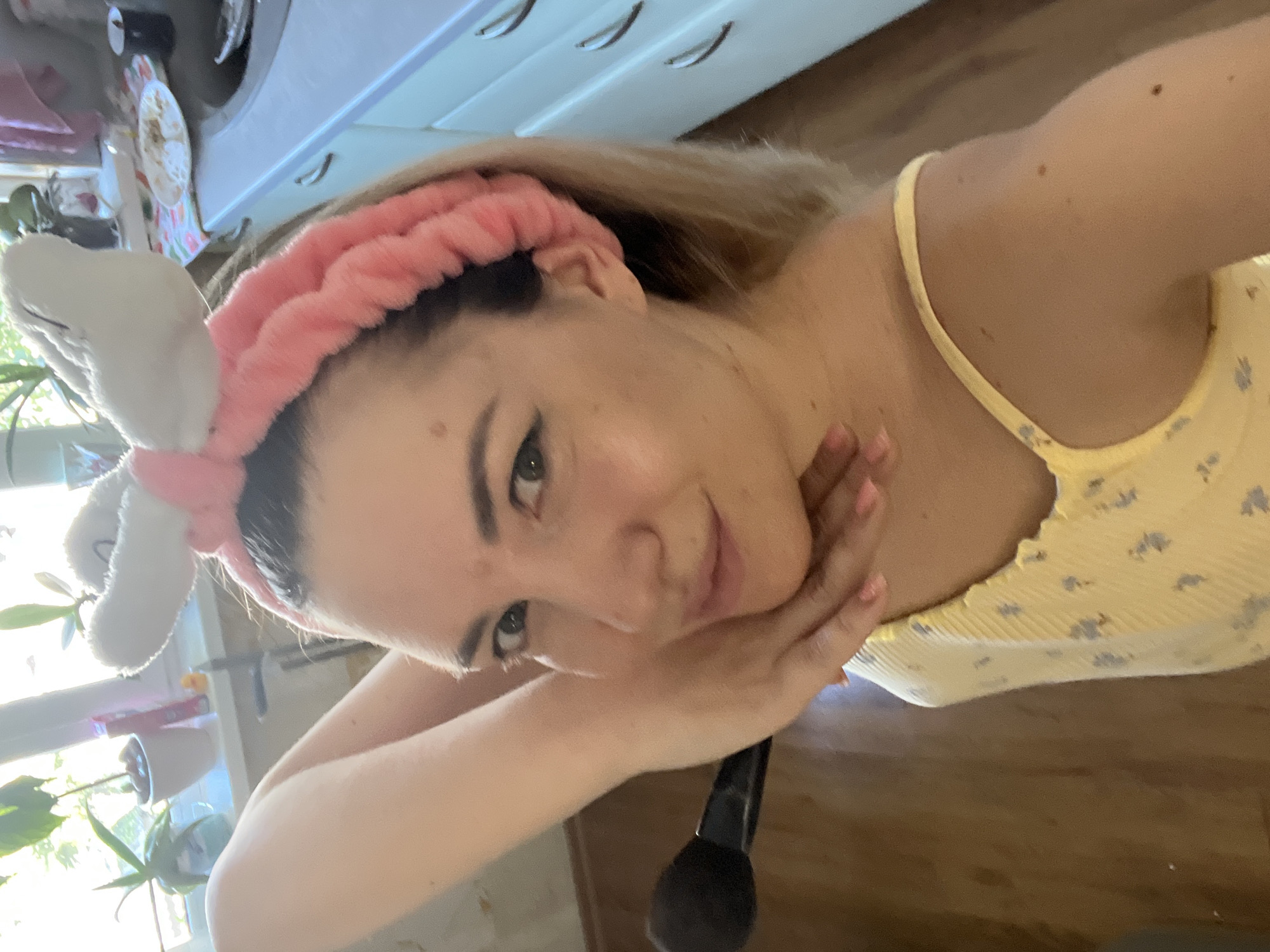 The media file is a selfie taken by a young woman with blonde hair and light skin. She is wearing a fluffy pink headband with white bunny ears and a yellow floral print top. Her face is angled towards the camera, with her chin resting on her hand. She has makeup on, including eyeliner and mascara. A black makeup brush is visible near her hand. The background appears to be a domestic setting, possibly a bathroom or bedroom, with cabinets and plants visible. The lighting suggests it is daytime. The overall mood seems relaxed and casual. There is no visible text in the image.