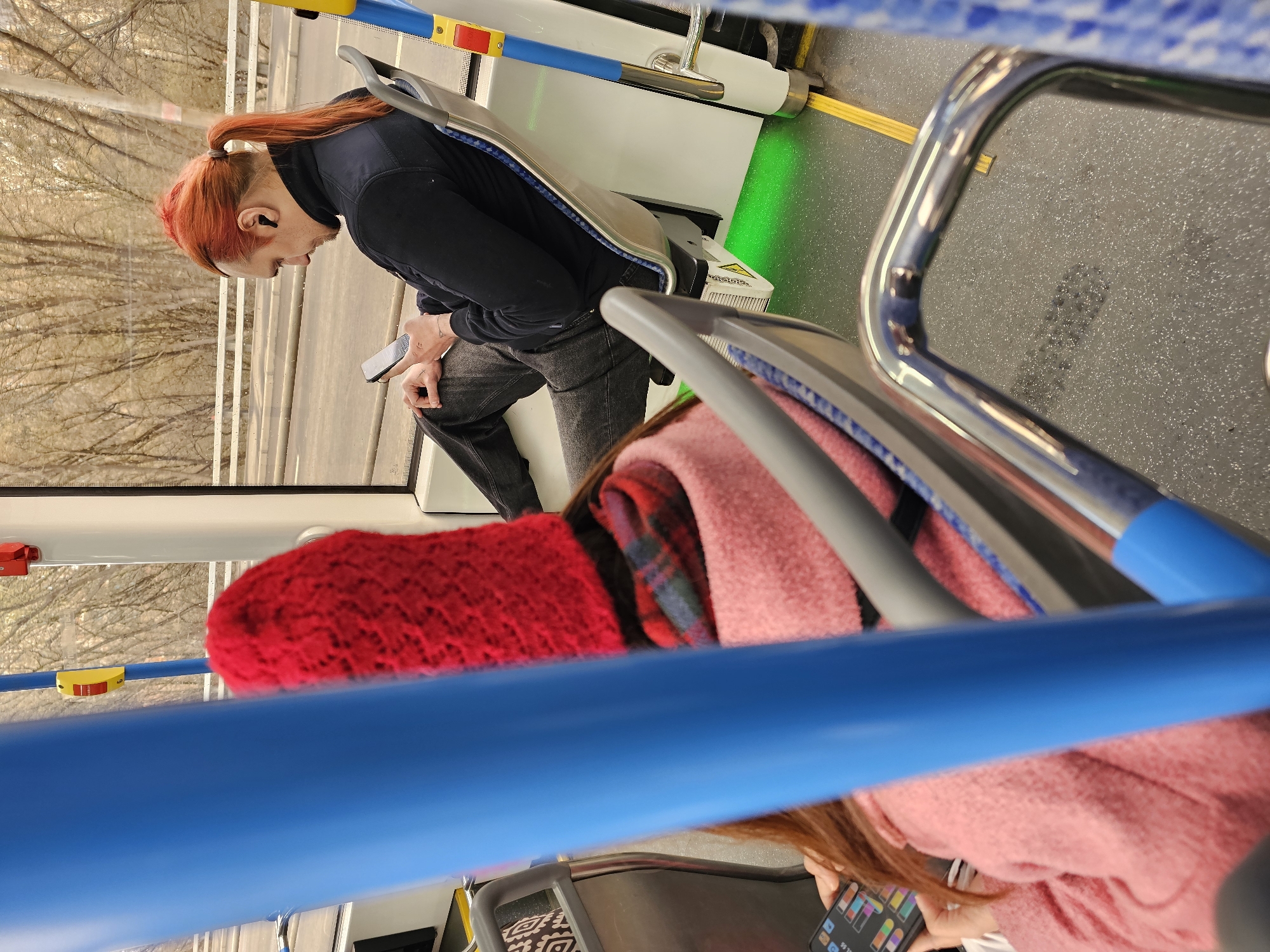 On a bus, a person with orange hair tied in a ponytail is looking at a smartphone in their hands. They are wearing a black jacket and gray jeans. Another person is sitting in the foreground, wrapped in a pink coat and a red scarf. A blue handrail is in the foreground. Through the bus window, trees without leaves can be seen. The location is Khimki, Russia.