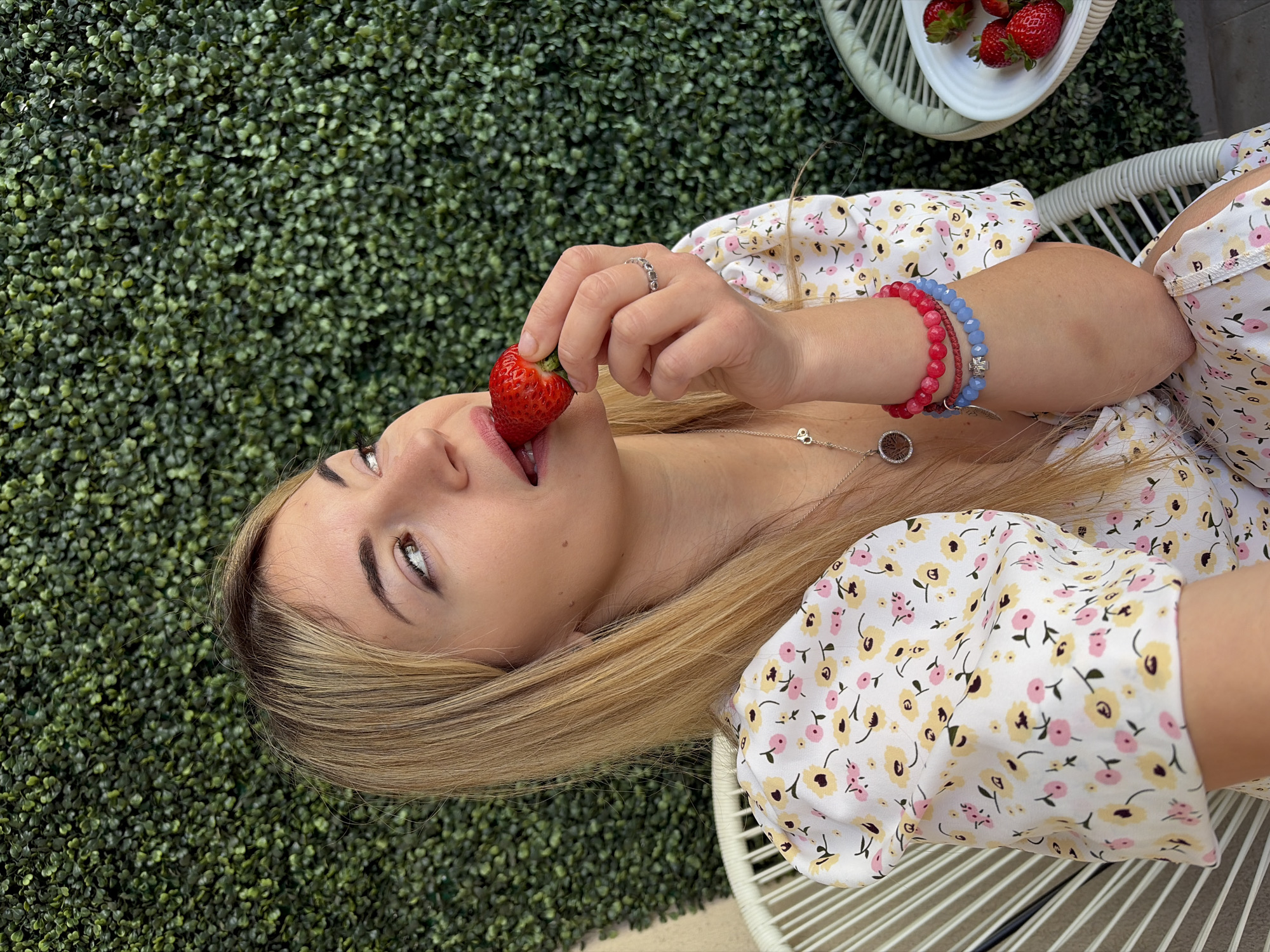 A woman with long blonde hair, wearing a white floral dress, is seen in an eye-level, candid shot, holding a strawberry to her lips. She is seated on a white, rounded chair with a latticed design. Her left hand, adorned with a ring, holds the strawberry. Her right wrist has two bracelets, one red and one blue. She is wearing a silver necklace. A small white plate with strawberries is visible in the upper right corner. The background is a wall of greenery. It is not possible to confirm that the location is in the city of Budva, Montenegro based solely on the visual data provided.