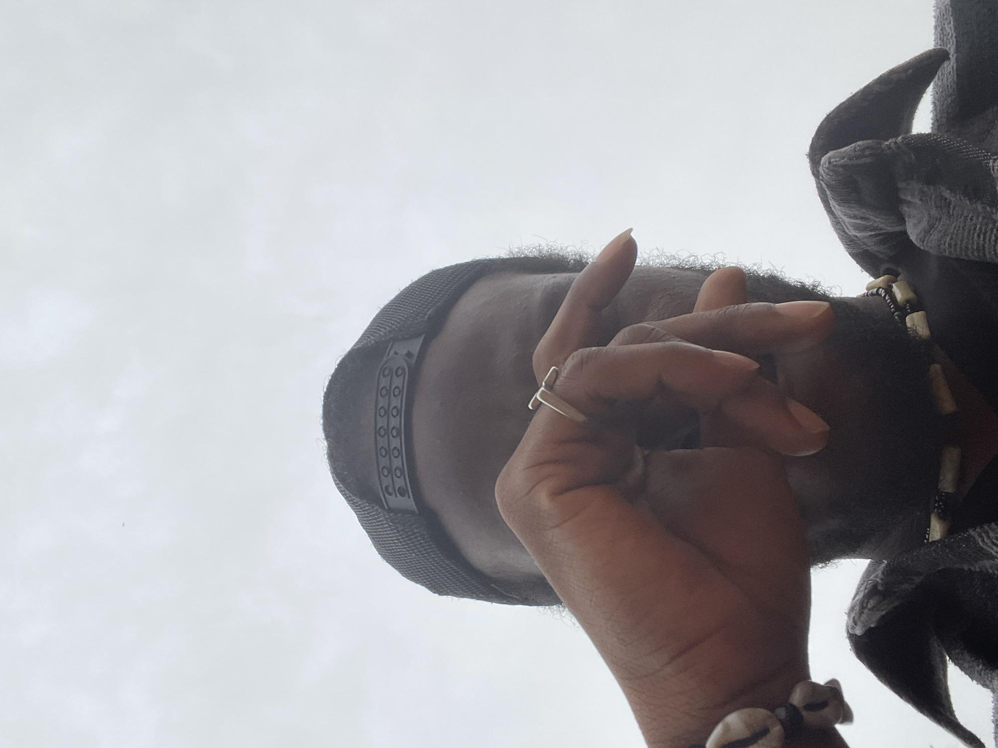 A low-angle shot of a person looking down towards the camera. The person is wearing a dark-colored cap with a decorative band. The person's right hand is raised, with the index finger extended, and a ring on the finger. A bracelet adorns the wrist. The person is wearing a patterned shirt and a necklace with small, light-colored objects. The background is an overcast sky. There is no evidence in the image to determine if the location is Jos, Nigeria.