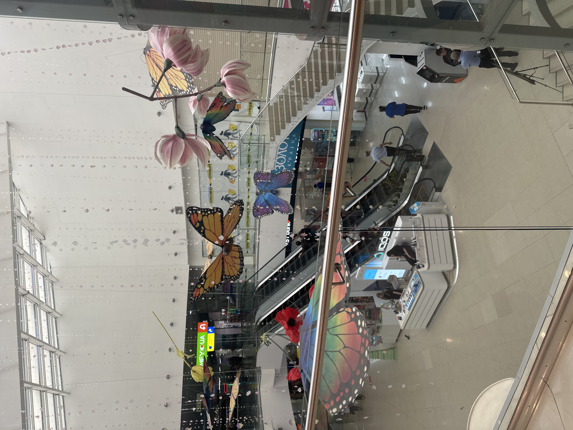 An indoor shot captures a multi-level shopping mall adorned with large, colorful butterfly decorations and delicate hanging strings of pale pink and white translucent shapes. A glass railing in the foreground partially obstructs the view of a lower level. On the lower level, escalators transport shoppers between floors. Several people are visible: two individuals walk up an escalator on the left, one person walks across the floor on the right, and two men in uniform stand near a large grey machine on the right, with one appearing to interact with it. Another man in uniform is seated on a bench. In the center of the lower level, a display counter with white shelving and products is manned by two women. The environment features a polished tile floor and various storefronts. The overall impression is a bright, spacious commercial interior decorated for a seasonal or promotional event.