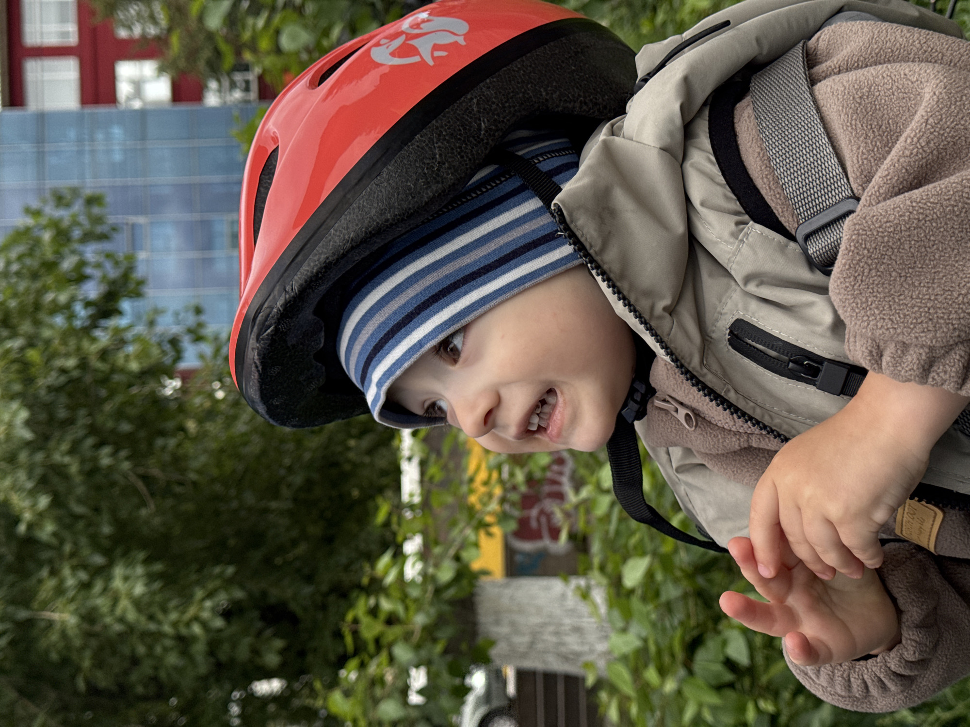 A close-up, tilted shot shows a young child wearing a red helmet and a striped beanie. The child has an excited expression with their mouth open, revealing teeth. They are also wearing a light tan jacket over a brown fuzzy garment. The child's hands are visible, one resting on the jacket and the other open, showing its palm. A black strap is attached to the helmet.
The background is out of focus, with green foliage and a building with blue glass windows and red accents. The image appears to be taken outdoors during the day. There is a small tag on the child's jacket with some unreadable text. The setting suggests an outdoor activity, possibly a bike ride or a walk, in Izhevsk, Russia. The child's expression conveys happiness and anticipation.