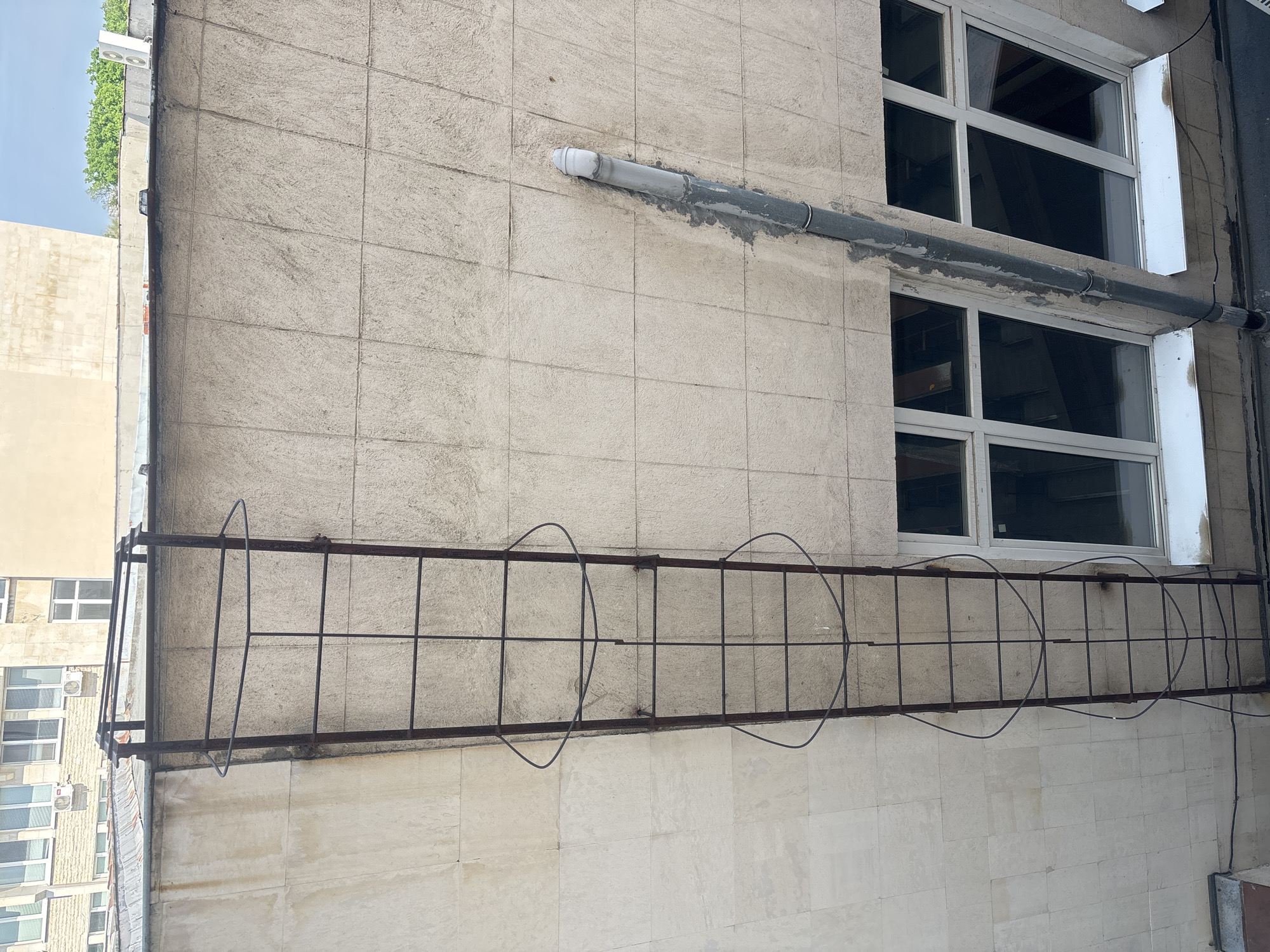 The image displays the exterior side of a building wall, predominantly tiled in a light beige or grey hue, characterized by a pattern of square tiles. Two horizontal rows of white-framed windows are visible, with their glass appearing dark, suggesting reflections or shadowed interiors.
A thick grey pipe runs horizontally across the tiled wall, positioned above the upper windows and partially secured with a rough grey cement-like material. Another thinner, dark pipe or cable extends vertically along the far right edge of the frame. Below the windows, a long, rusty, dark metal grid structure is attached to the wall. This structure, possibly a rudimentary balcony or security barrier, is composed of horizontal and vertical bars and features two distinctive large, bent, circular wire elements on its upper edge.
To the left, a portion of an adjacent building is visible, featuring numerous windows and what appear to be air conditioning units. Green foliage is noticeable at the top left, set against a bright, clear blue sky. The scene is captured in daylight, with no discernible human activity or visible text. The overall impression is an urban facade, suggesting a side or rear view of a building in a city like Plovdiv, Bulgaria, with functional elements and signs of weathering.