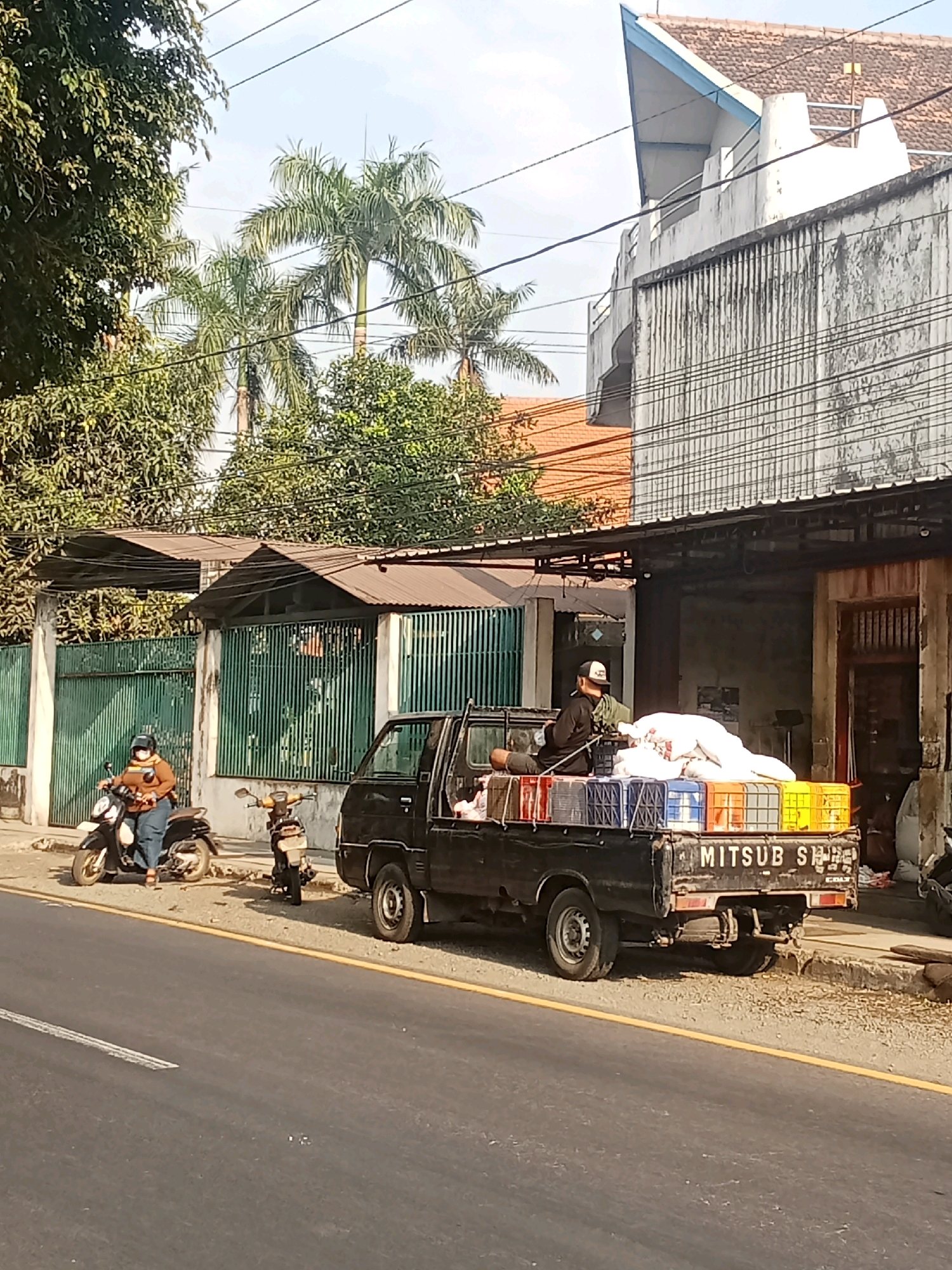 The image depicts a street scene in Kaliwungu, Indonesia, on a sunny day. A black Mitsubishi pickup truck is parked on the side of the road, loaded with white sacks and colorful plastic crates. A person wearing a black cap and shirt is seated on top of the load in the truck bed. To the left of the truck, a person wearing a brown jacket and mask is riding a black scooter, with another parked scooter next to it. In the background, there are buildings with tiled roofs and palm trees, suggesting a tropical environment. The road has a yellow dividing line. The scene captures a moment of daily life, likely related to transport or commerce in the area.