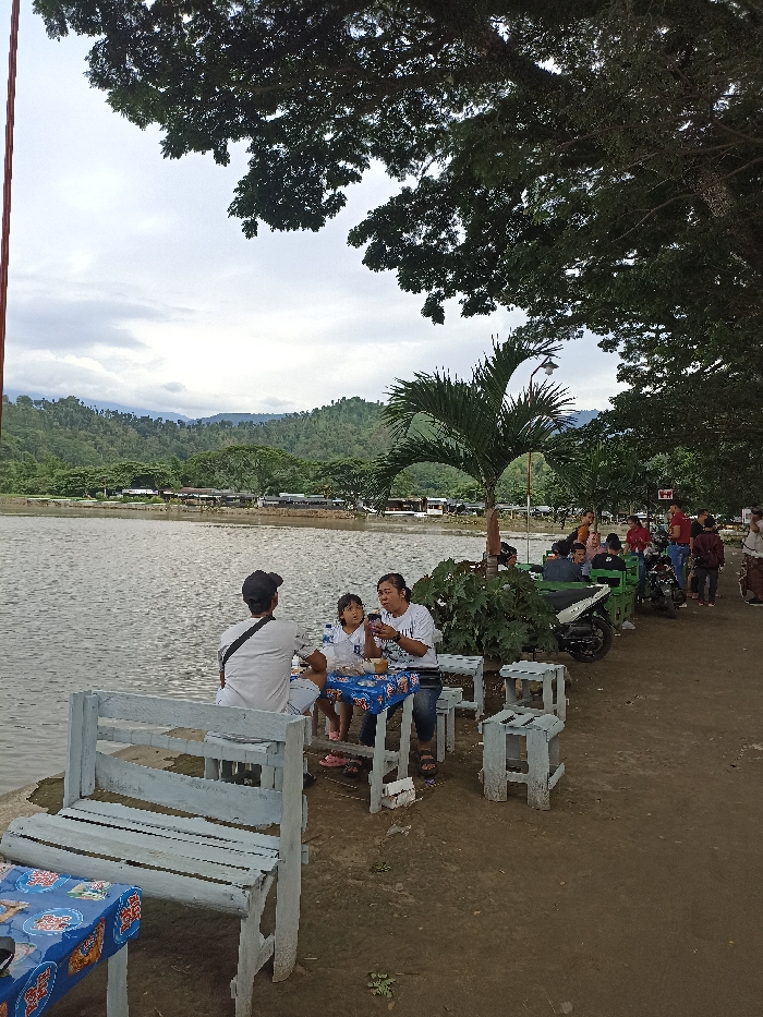 The image displays an outdoor scene by a body of water in Kepung, Indonesia, likely a lake or large pond, under an overcast sky. In the foreground, a family of three – a man, a woman, and a young child – is seated at a small, light-colored wooden table. The table is draped with a blue and white patterned tablecloth, on which food containers and drinks are placed. The woman is holding a smartphone, with the child looking towards it. The man, seen from behind wearing a white shirt and a dark cap, faces the water.
Behind the family, the calm water stretches towards a distant shore lined with various buildings and structures, backed by lush, forested hills and mountains. The sky is predominantly cloudy, indicating an overcast day or possibly late afternoon.
To the right of the seated family, a dirt path runs parallel to the water. Further along this path, more people are visible, some standing and socializing, while others are near parked motorbikes. A slender palm tree stands prominently, adorned with a street light, and large, dense-leafed trees frame the upper and right portions of the image, providing ample shade. The seating consists of simple, light-colored wooden benches and stools. The overall atmosphere is relaxed, suggesting a casual recreational or dining spot. There is no clearly legible text visible on signs or clothing.