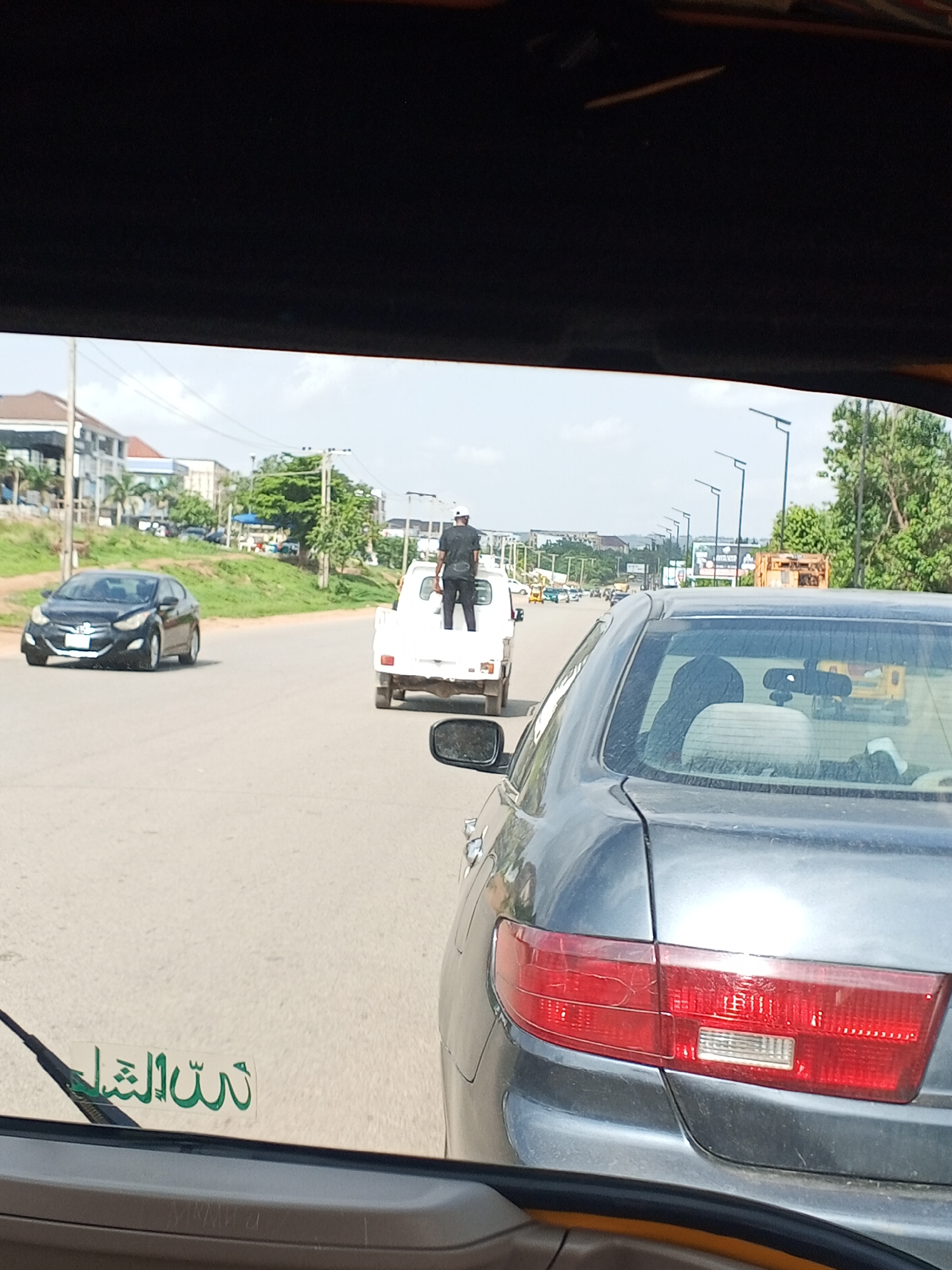 The photo, taken from inside a vehicle in Galadimawa Village, Nigeria, shows a street scene with multiple cars on the road. In the foreground, a gray car’s rear is visible. Ahead of it, a white pickup truck has a person standing in its bed. A black car is in the leftmost lane, moving in the same direction as the other vehicles. The background includes buildings, trees, and street lights under a slightly overcast sky. The view is framed by the dark interior of the vehicle from which the photo was taken.