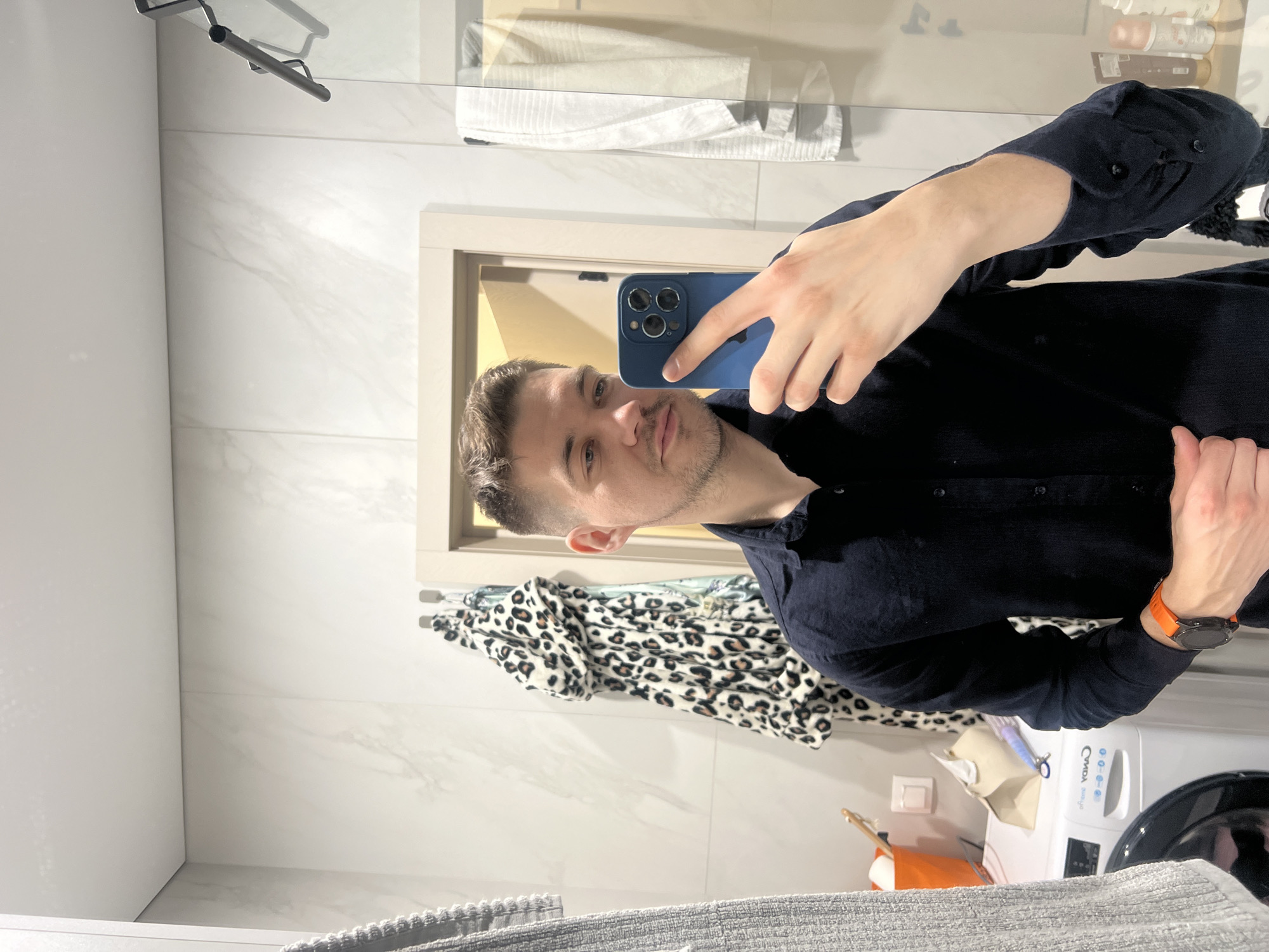 A young adult male with short brown hair and light skin is depicted in a bathroom, taking a mirror selfie. He wears a dark blue long-sleeved shirt and an orange-strapped smartwatch on his left wrist. His right hand holds a blue smartphone with a prominent triple-lens camera system, aimed at the mirror. The background features light-colored walls tiled with a subtle marble pattern and a white ceiling.
To the left, a white Candy brand washing machine is visible. Above it, on a surface, are an orange bucket containing cleaning brushes and toilet paper rolls, a light-colored tissue box, and a purple-handled grooming tool. A white electrical outlet with a switch is mounted on the wall near these items. A light-colored wooden door frame is partially visible behind the subject's head. On the wall to the right of the door frame, two silver hooks hold a white and black leopard-print robe and another patterned fabric item. Further right, a white towel hangs over a metal bar, which appears to be part of a shower enclosure. Various toiletry bottles are visible on a shelf reflected in the mirror behind the male's right shoulder. This scene occurs within a private residence in Kazan, Russia.