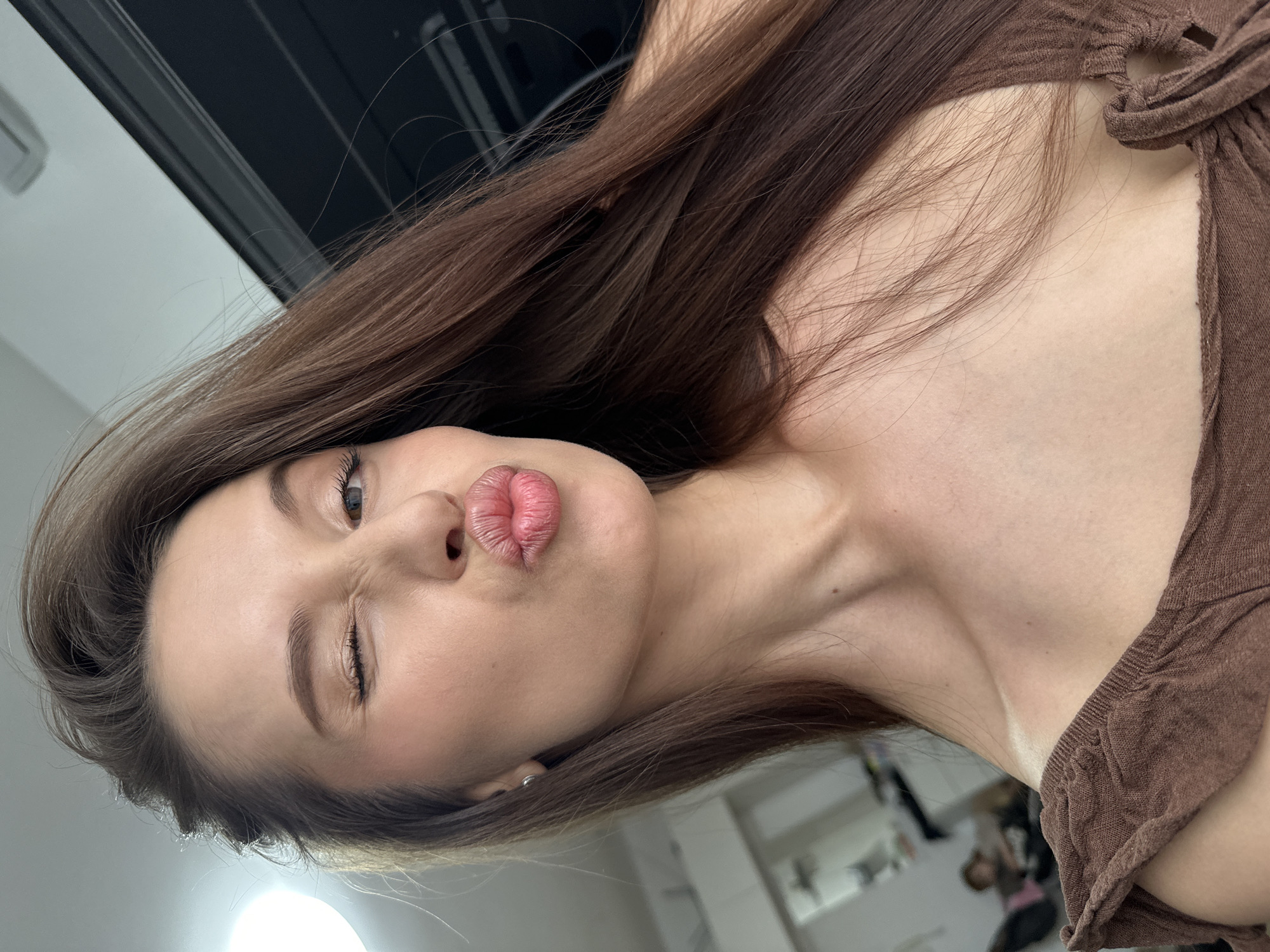 A woman with long brown hair and fair skin is looking at the camera. She is positioned at an angle, with her head tilted. She is pouting her lips and winking one eye. She is wearing a brown garment. The background is blurred, with glimpses of what appears to be a room interior with light and dark elements. There is no indication this picture was taken in the city of Yekaterinburg, Russia.