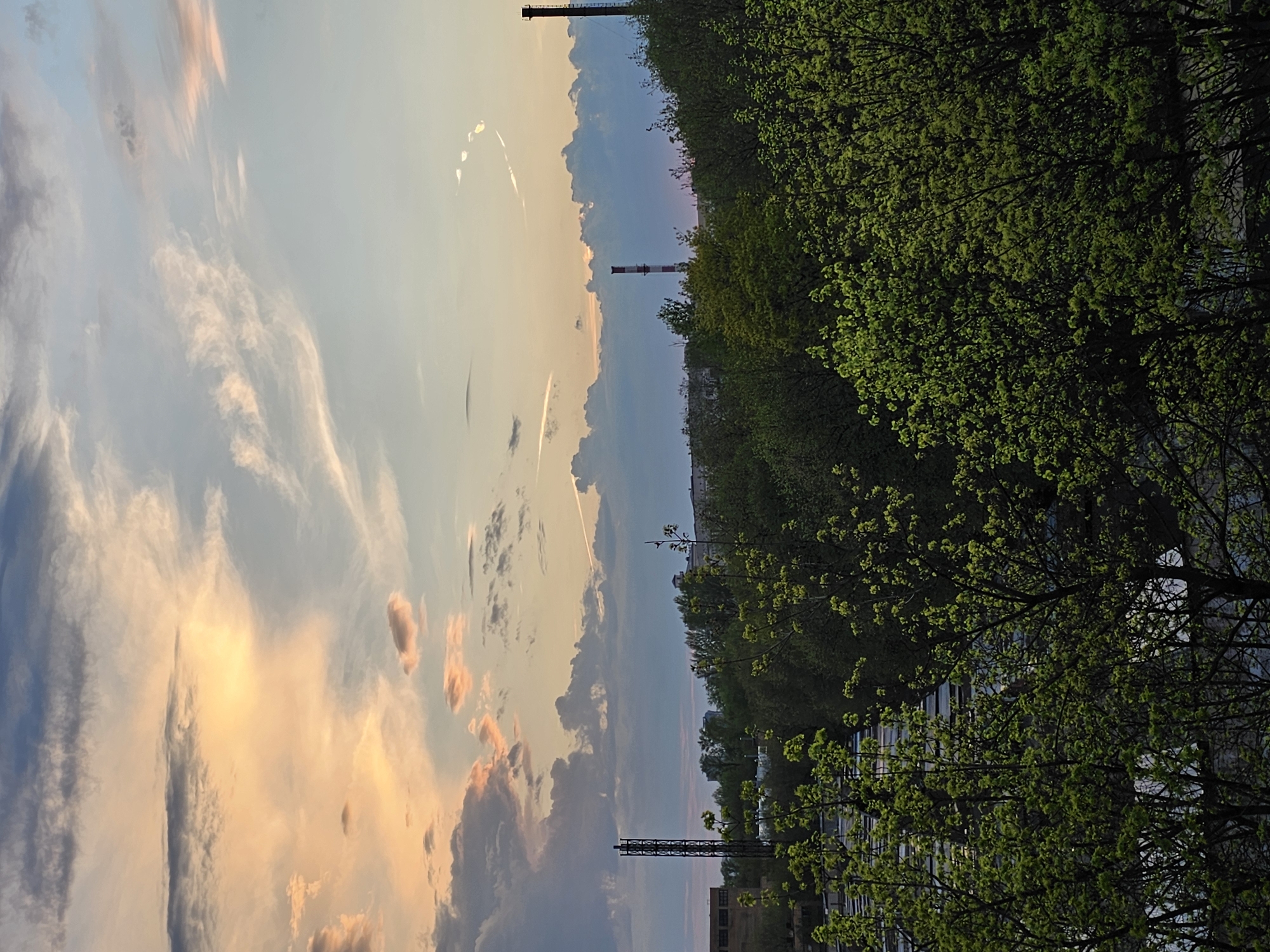 A landscape-oriented photograph shows a sky filled with clouds and two industrial chimneys. The sky shows hues of orange, pink, and blue at sunset. To the right, a thick row of trees with green leaves partially obscures a view of a city street. Buildings and lampposts are visible through the trees. The photograph is oriented with the ground at the top. The chimneys are characteristic of Moscow’s thermal power plants.