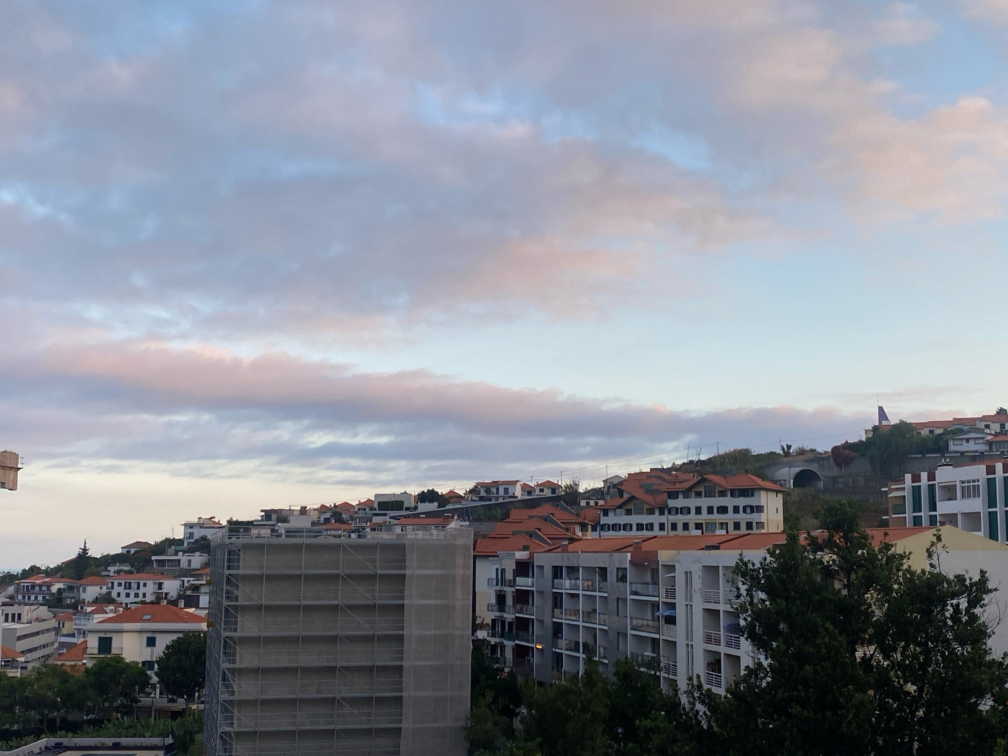 This image depicts a view of the city of Funchal, Portugal, likely from an elevated position, showcasing a hillside covered in buildings and vegetation under a cloudy sky. In the foreground, a large building under construction, wrapped in scaffolding and netting, dominates the left side of the frame. To its right, several multi-story apartment buildings with white facades and dark-trimmed balconies are visible. Further up the hillside, a dense collection of residential houses with red-tiled roofs are nestled amongst trees. The sky above is filled with soft, wispy clouds tinged with pink and purple, suggesting either sunrise or sunset. The overall scene portrays a vibrant, densely populated urban environment on a hilly terrain.