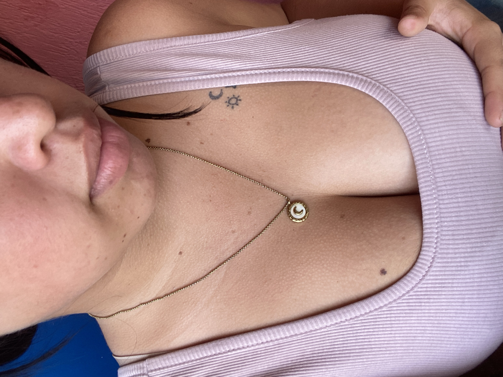 A close-up, angled shot focuses on a person's chest and face. The individual is wearing a light purple ribbed tank top. A gold necklace with a small, round pendant rests against their skin. A tattoo depicting a crescent moon and a sun is visible near their collarbone. Part of their face, including their nose and lips, is in view, as is some of their hair. The image does not provide any information about the location in the city of Juan Griego, Venezuela.