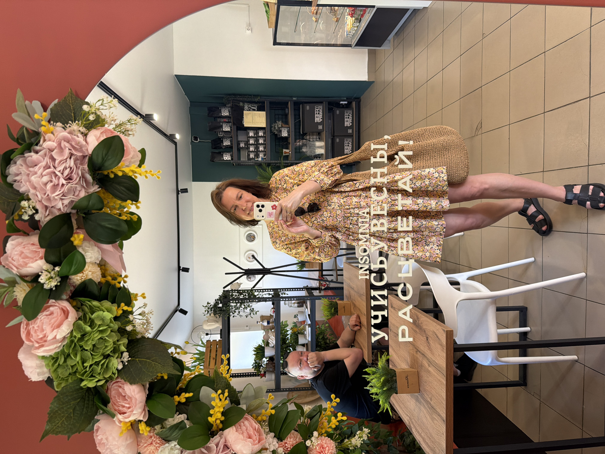 A woman in a floral dress takes a selfie in a round mirror decorated with flowers. The reflection shows a man sitting at a wooden table, various shelves with items, and white chairs on a tiled floor. The writing "INSOMNIA" and "учисьувесиы, РАСЦВЕТАЙ!" is visible in front of the mirror. The location is in Chaikovsky, Russia.