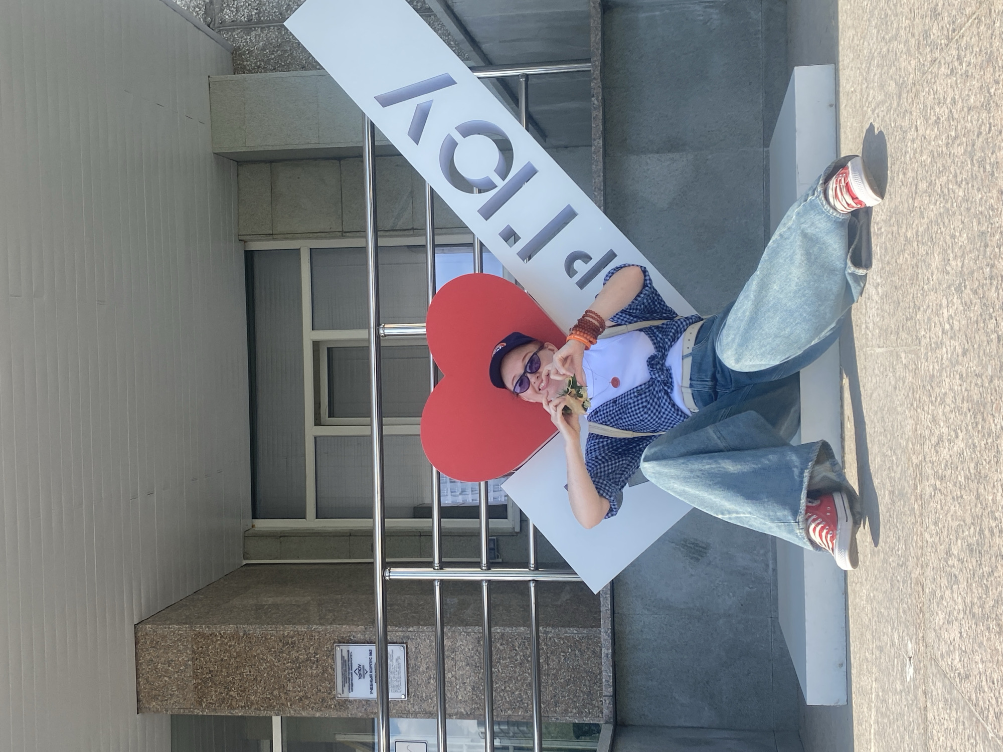 A person lies on a white sign with the word "UrFU" vertically oriented, overlaid with a red heart, in front of a building. The person wears a blue plaid shirt over a white top, light blue jeans, and red shoes. They have a dark baseball cap, sunglasses, and multiple bracelets. They hold something near their mouth. The location is in front of the Ural Federal University (UrFU) in Yekaterinburg, Russia.
