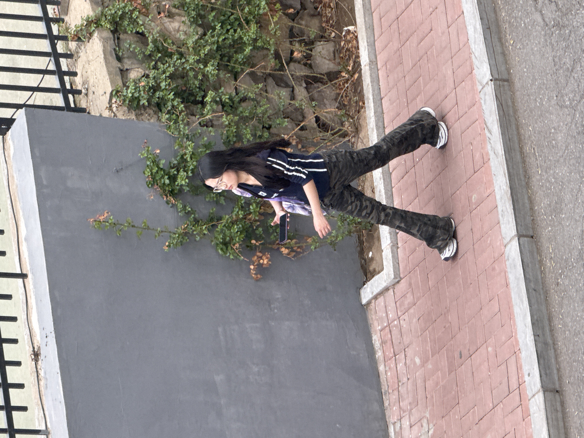 A young woman with long dark hair is walking on a brick sidewalk. She is wearing a dark jacket with white stripes, camouflage pants, and white sneakers. She is holding a cell phone in her hand. To her left is a gray wall with a green vine growing on it. The location is Zhongtun, China.