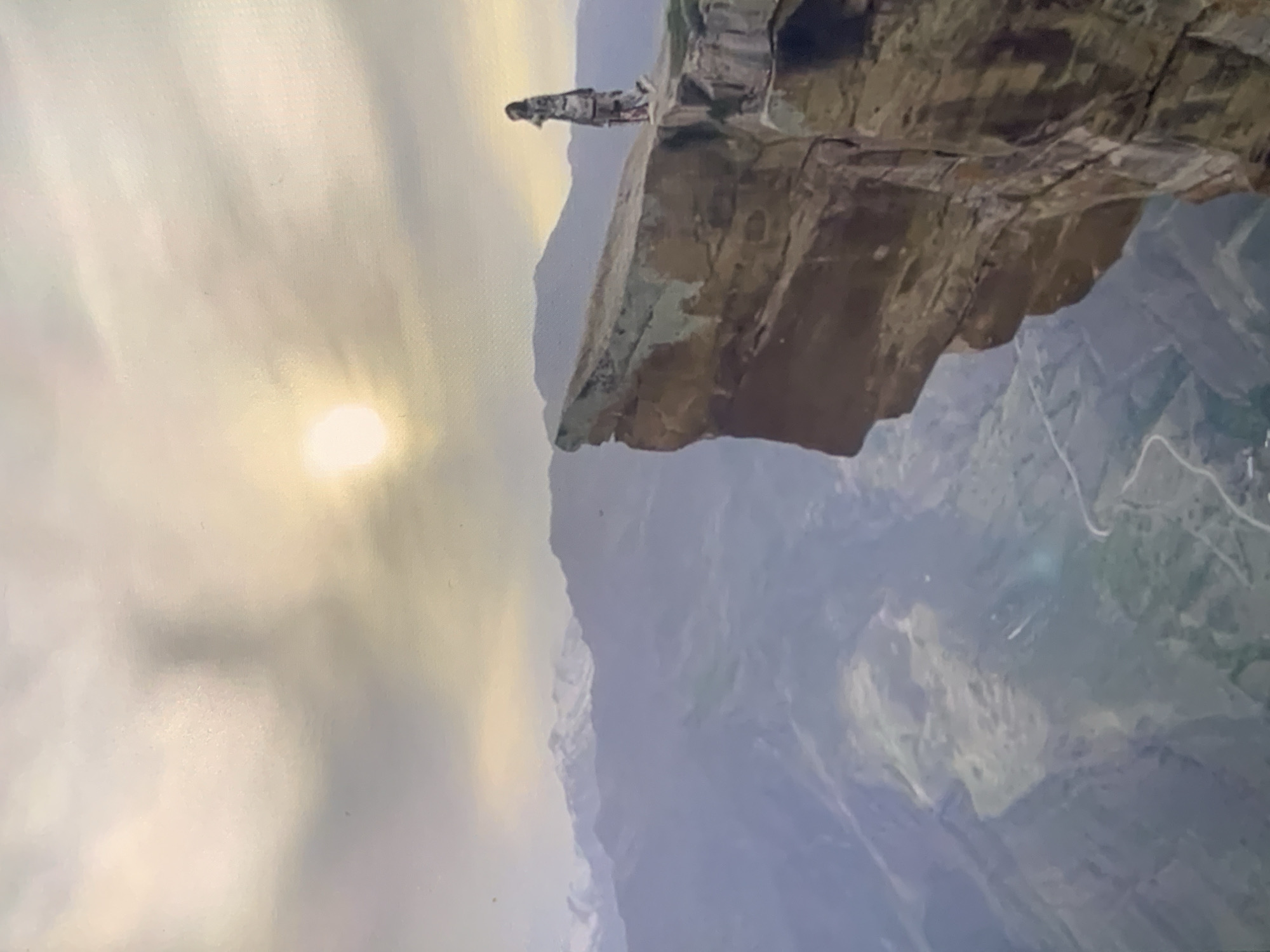 The image depicts a vast mountainous landscape under a cloudy sky with the sun partially visible, casting a warm glow. In the foreground, a person stands on the edge of a steep, rocky cliff. The individual is wearing a patterned outfit and appears to be looking out at the vista. The cliff face is sheer and rugged, with distinct layers of rock visible. Below the cliff, a valley unfolds, revealing more mountains in the distance, some with snow-capped peaks. A winding road is visible in the valley floor. The scene conveys a sense of awe and solitude. This location is not in the city of Stavropol, Russia, as Stavropol is a city and not a mountainous region. The image is of a natural, rugged mountain environment.