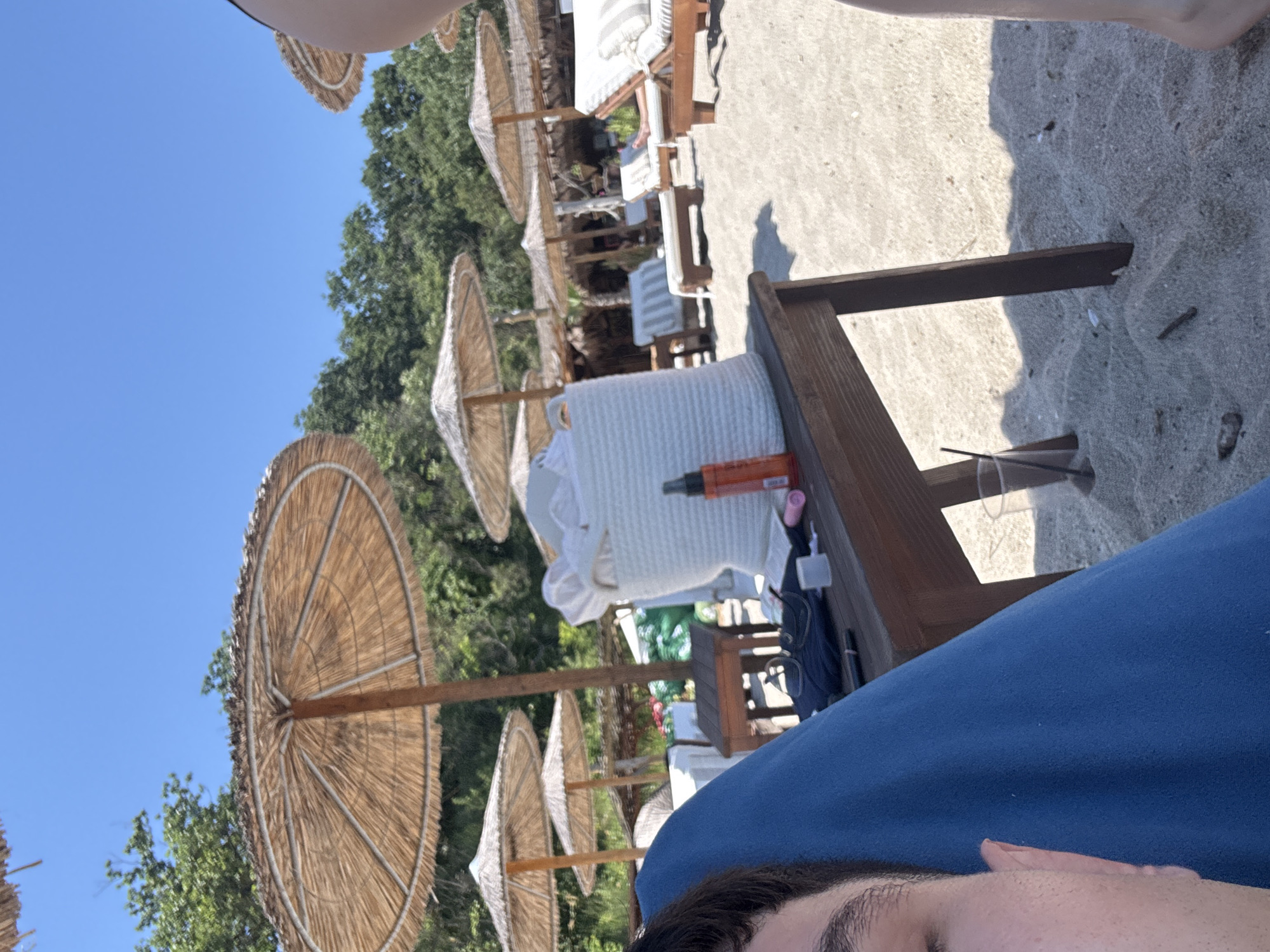 The image depicts a beach scene on a sunny day, likely in Varna, Bulgaria, given the mention of the location. Several thatched umbrellas are set up on the sandy shore, providing shade for lounge chairs arranged on the beach. In the foreground, a person's face is partially visible, along with a blue towel they are lying on. A wooden table is positioned nearby, holding various items, including a white woven basket, a bottle of sunscreen, glasses, and a cup with a straw. In the background, lush green trees form a backdrop to the beach. The overall impression is one of a relaxing day at the seaside.