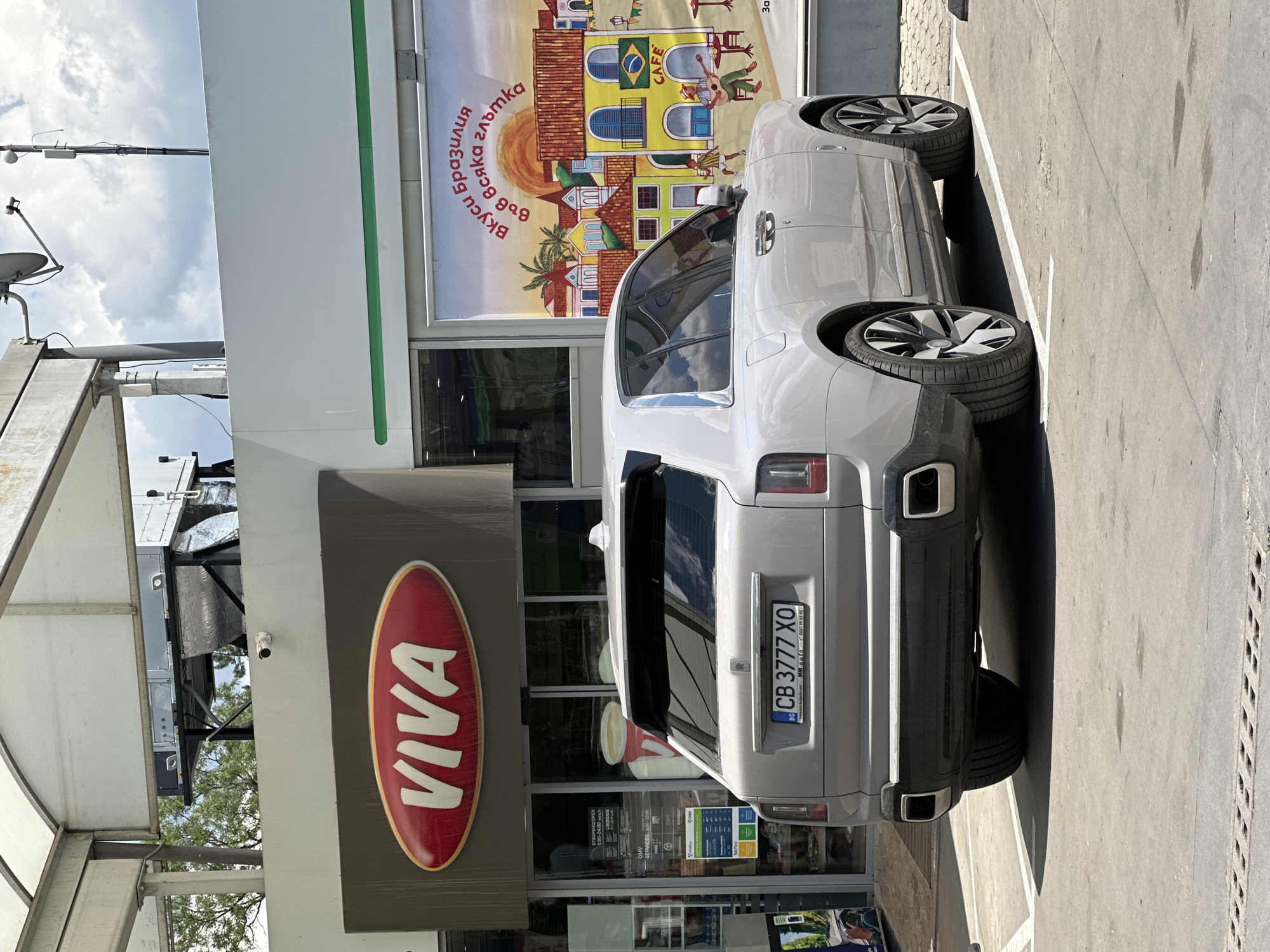 A light gray Rolls-Royce Cullinan SUV is parked in a lot in front of a Viva gas station. The car is facing away from the camera, with its rear bumper and license plate visible. The license plate reads "CB 3777 XO" and has a Bulgarian flag on the left. The Viva gas station has a large red oval logo with "VIVA" written in white letters. To the right of the gas station entrance is a large advertisement with a colorful depiction of a Brazilian village. The scene is set on a sunny day, with clouds in the sky. The background includes trees and a portion of the gas station's canopy. The location is the Viva gas station on the road from Veliko Tarnovo to Gabrovo, near the village of Chirpan. There are no people visible in the image.