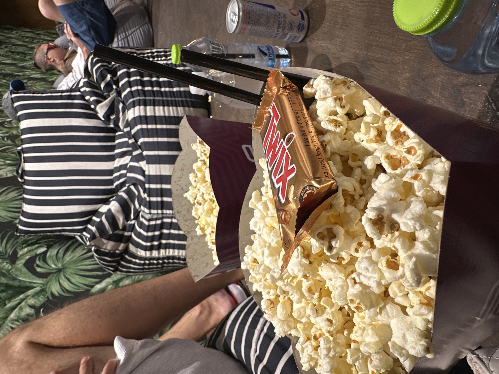 This image depicts a casual gathering, likely a movie night at home or a private cinema, featuring two large containers of popcorn. One container, in the foreground, is overflowing with popcorn and has a partially unwrapped Snickers bar placed on top. Two black straws are inserted into this container. Another smaller container of popcorn is visible in the background. In the background, a person is seated on a striped beanbag chair, looking at a laptop. A bottle of water with a green cap and a can are on the table. The scene appears to be set in a living room with a tropical-themed wallpaper. While the image does not provide explicit visual cues to pinpoint the exact location in Netanya, Israel, the presence of Hebrew text on the Snickers wrapper suggests it was taken in Israel.