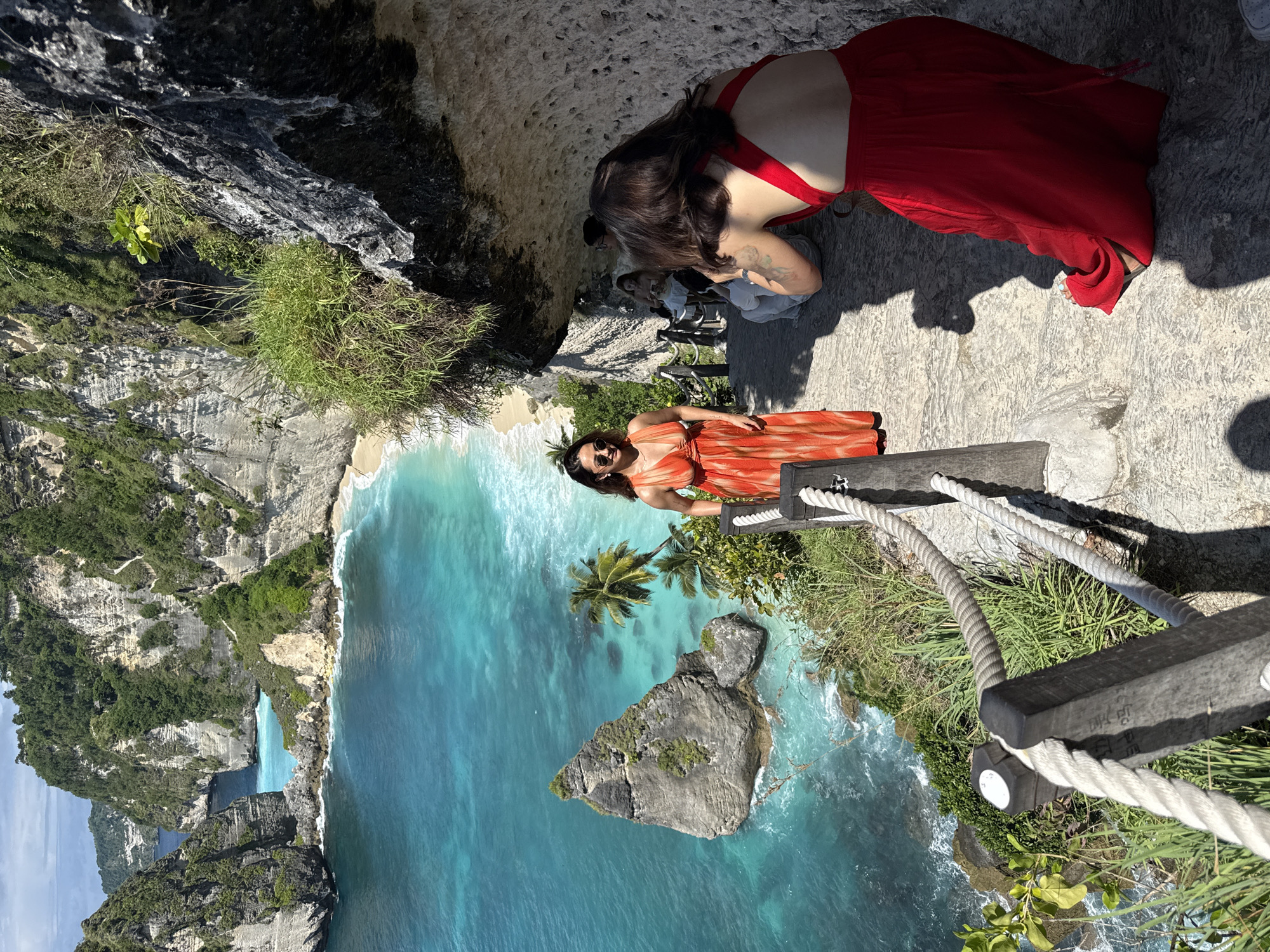 The image depicts a scenic view in Pejukutan, Indonesia, featuring a woman in an orange dress standing on a cliff edge overlooking a vibrant turquoise ocean. Beside her, a wooden rope railing provides a safety barrier. Further back on the cliff, another woman in a red dress is bending down, seemingly interacting with a photographer who is taking a picture. The cliff face is rugged with green vegetation. Below, a sandy beach meets the clear water, and a large rock formation with palm trees stands prominently in the sea. In the distance, other cliffs and ocean expanses can be seen. The lighting suggests it is a sunny day.