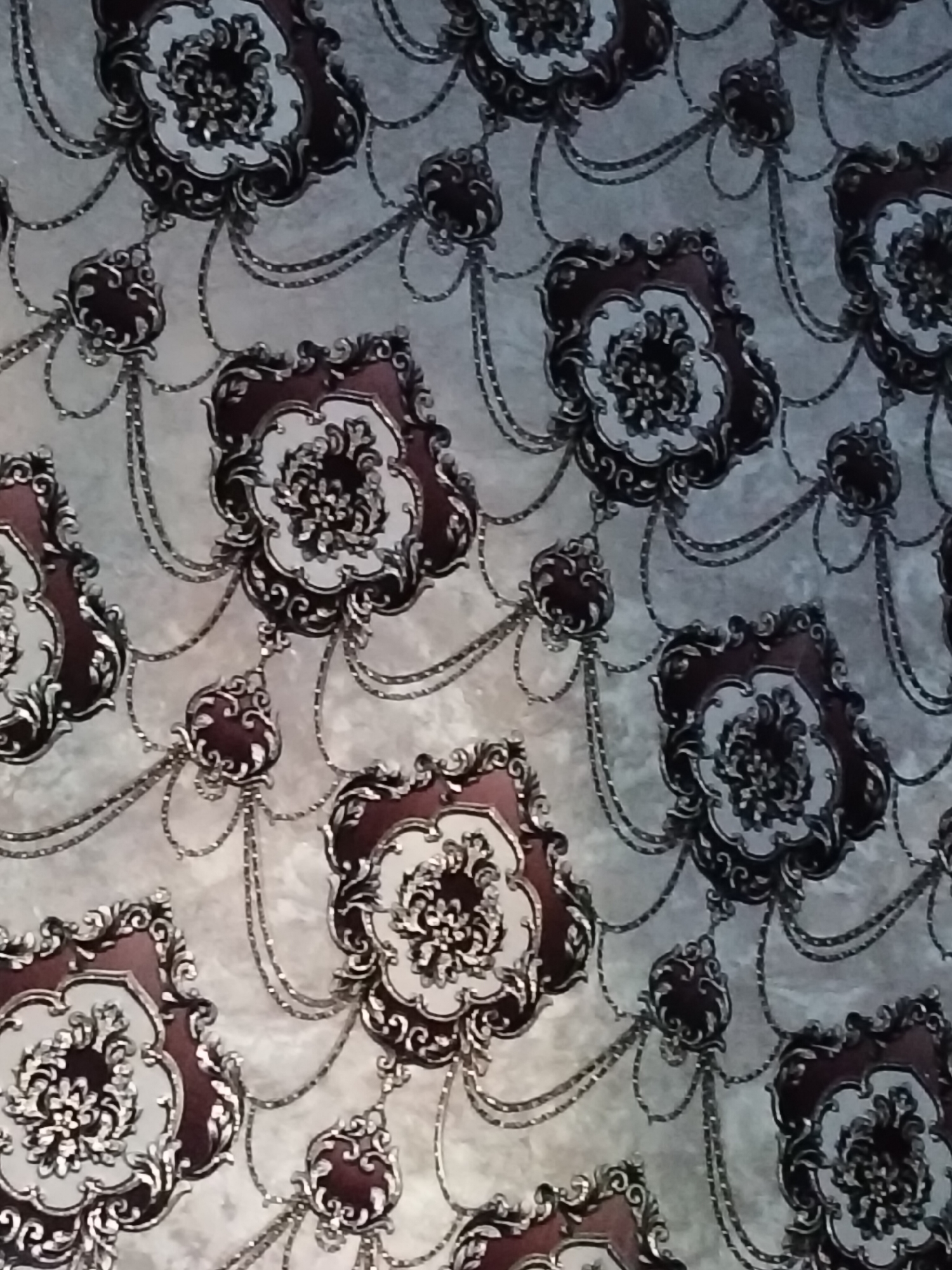 The image displays a close-up view of a patterned wallpaper with a baroque or rococo design. The pattern features ornate, floral motifs in dark red and white, outlined with black and silver accents. These motifs are interconnected by decorative chains and dangling elements, creating a sense of cascading ornamentation. The background of the wallpaper appears to be a textured, light gray or off-white surface, possibly resembling marble or silk. The lighting is somewhat dim, casting subtle shadows that accentuate the three-dimensional feel of the embossed pattern. There are no people or overt signs of a specific location within a city like Kogon City, Uzbekistan visible in this image; it solely focuses on the texture and design of the wallpaper itself.