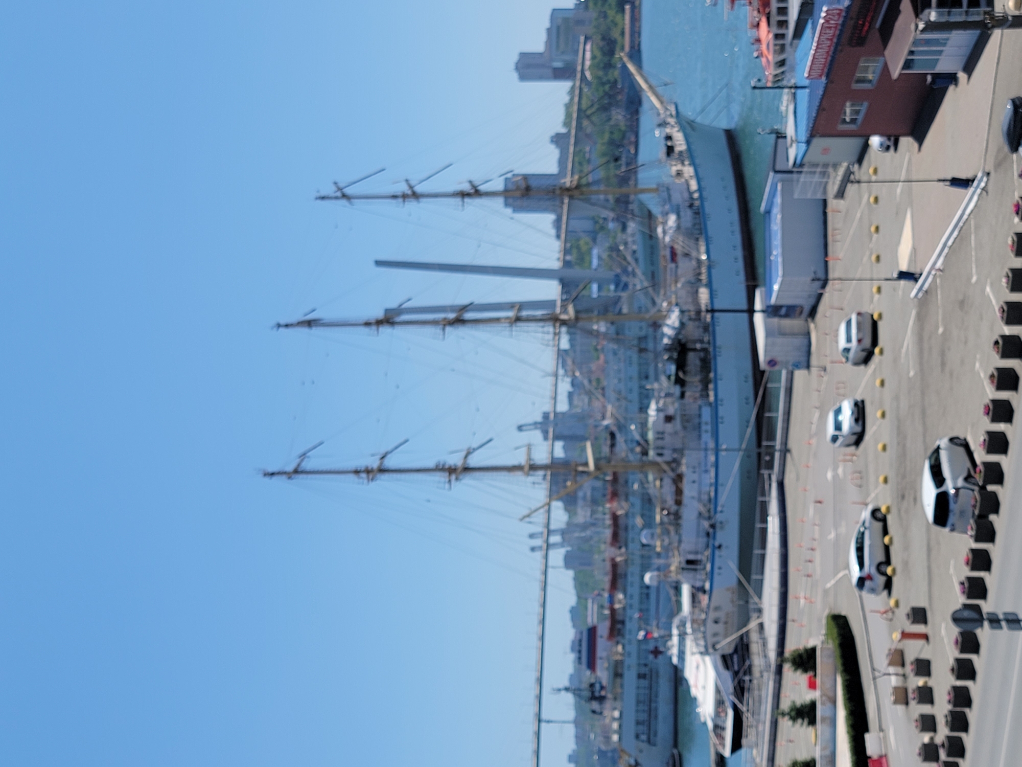 The image shows a large sailing ship docked in a harbor, next to a pier with a building. There are several cars parked along the pier, and the ship's masts stretch high into the blue sky. The location is in an unknown city. There are buildings visible in the background on the other side of the harbor.