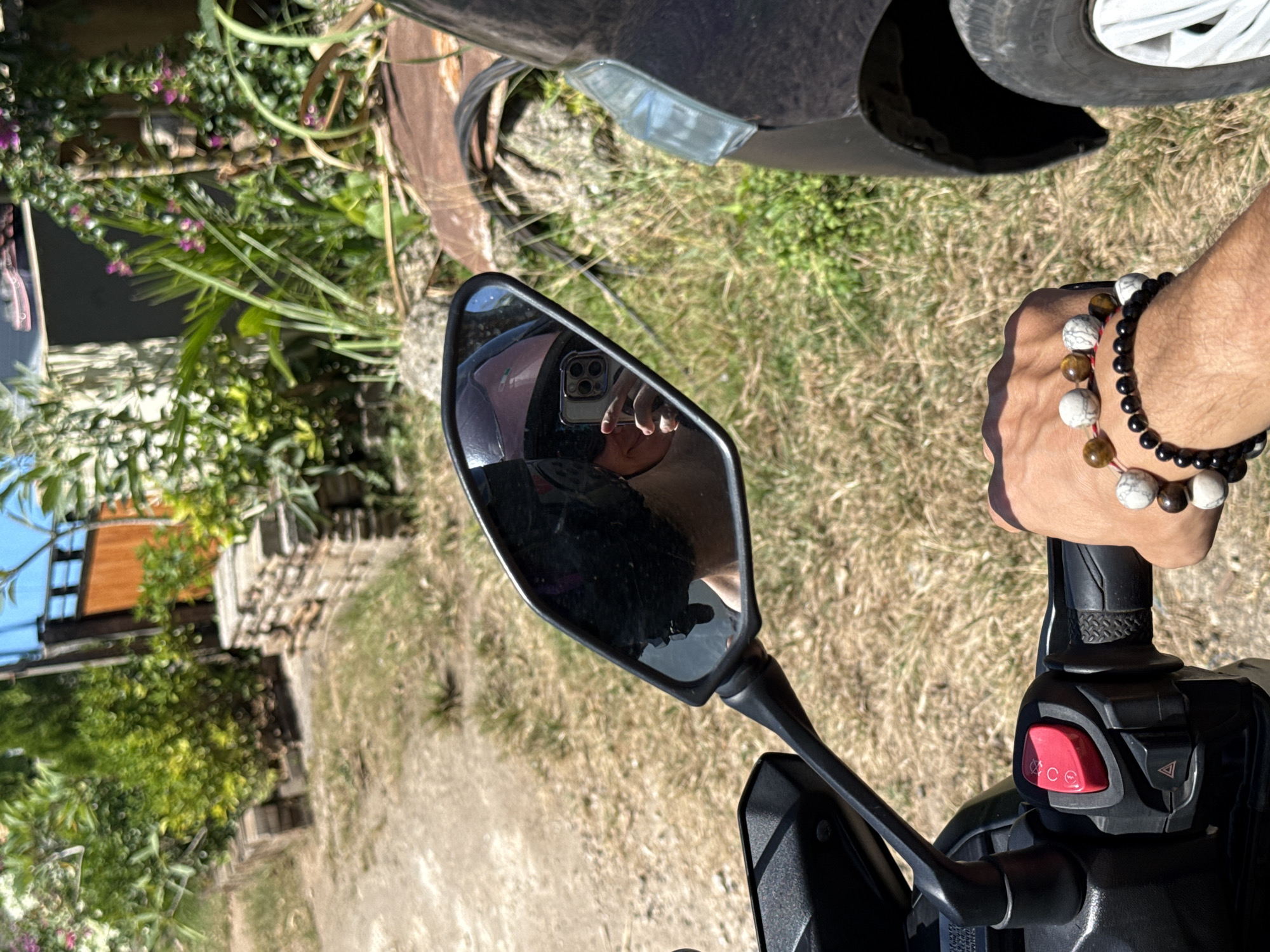 A first-person perspective shot shows a hand wearing beaded bracelets, resting on the handlebar of a dark-colored motorcycle in Kuta Selatan, Indonesia. The motorcycle's red start/stop button is visible. In the motorcycle's side mirror, a person wearing a helmet is reflected, holding a smartphone to take a photo. The background consists of dry grass, a dirt path, and lush green foliage with some flowering plants. A portion of a dark vehicle is visible to the right of the motorcycle.