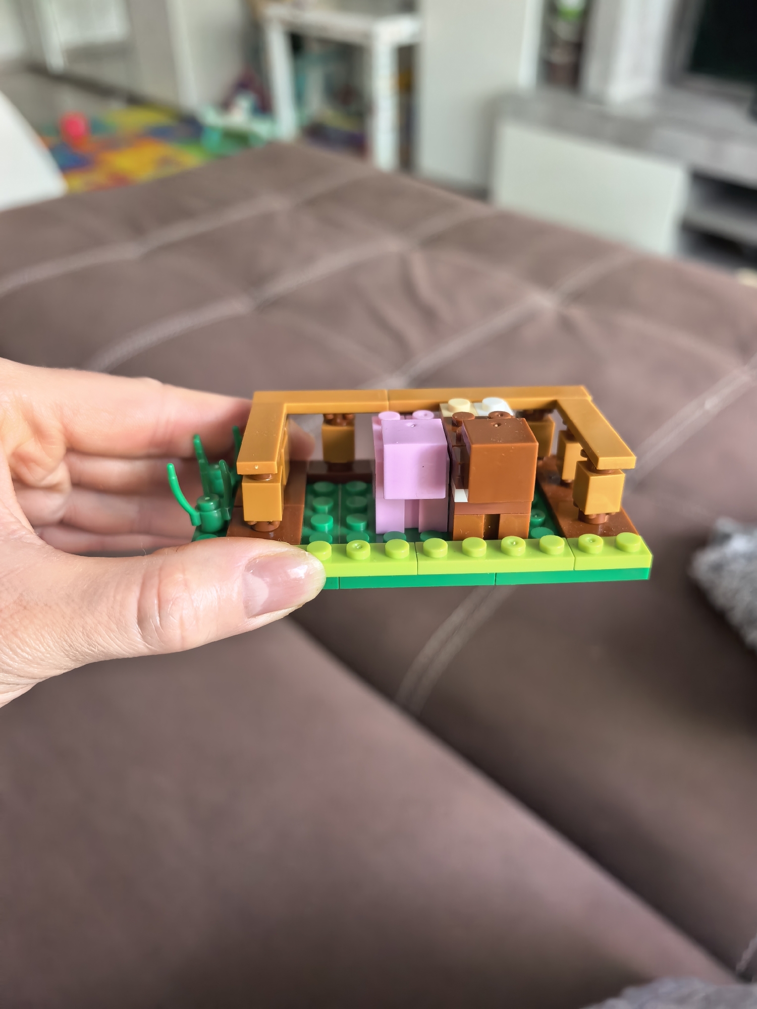 A person holds a Lego model of a Minecraft pig pen. The pen contains a pink Lego pig and a brown Lego cow. The base of the pen is green Lego bricks and brown Lego bricks. The pen has a brown Lego frame. The Lego model is held over a brown quilted surface. In the blurred background are various pieces of furniture. The location appears to be inside a house. There is no indication this image was taken in Sterlitamak, Russia.
