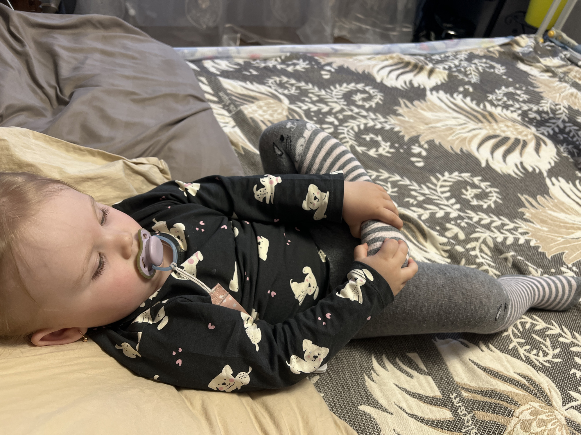 A young child is depicted lying on their left side on a bed in the city of Leninakan, Russia. The child, with closed eyes, wears a black long-sleeved shirt patterned with white cartoon animals and pink hearts, grey leggings, and grey and pink striped socks. A purple and light blue pacifier is positioned in the child's mouth. The child's right hand grasps their bent left leg. The bed features a light beige sheet under the child's head and a dark grey patterned blanket with light beige and white feather/leaf motifs. A small gold-colored earring is visible in the child's right earlobe.