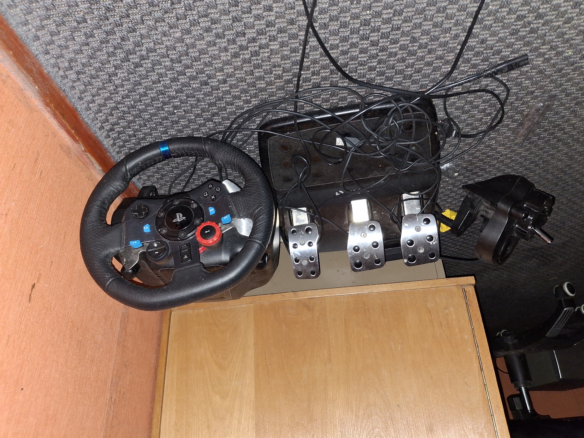 The image shows a gaming steering wheel and pedal set on a carpeted floor in a room with wooden furniture and an orange wall. The steering wheel is black leather with red and blue accents and various buttons. The pedal set consists of three metallic pedals on a black base, connected by wires. A black shifter is also visible on the left side of the image, with its wires tangled around the pedals. The scene appears to be a casual setting, possibly a home or office. The image does not provide any specific details about the city of Ogre, Latvia.