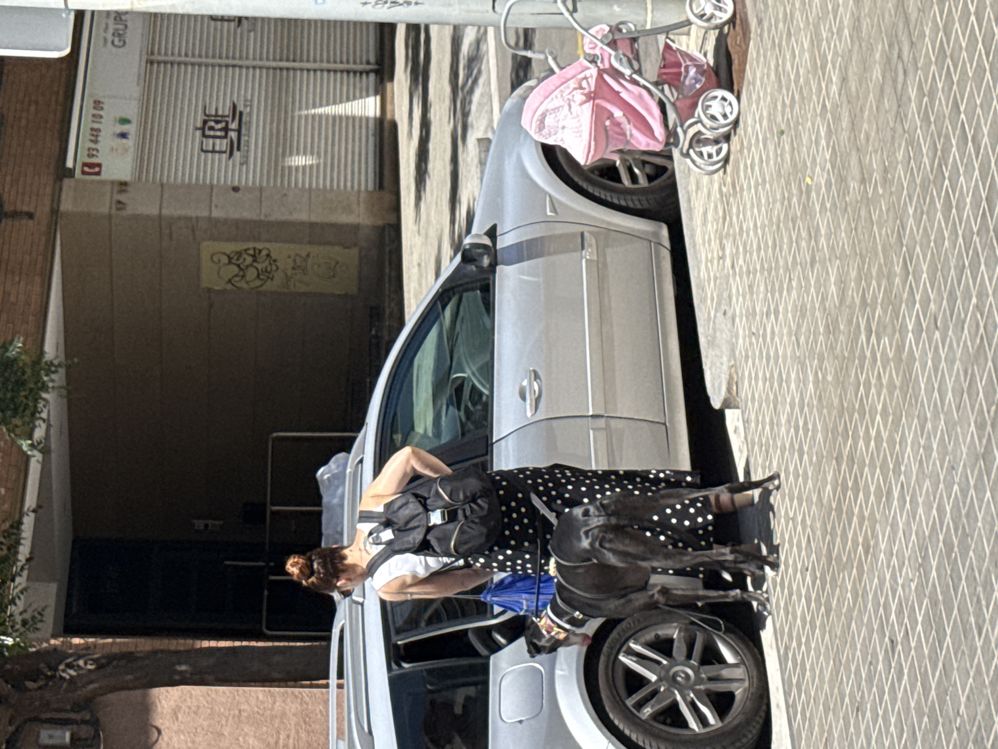 A medium shot shows a woman with her hair in a bun, wearing a white tank top and a black and white polka dot skirt, standing next to an open car door. She is holding a blue bag and a leash attached to a black dog that is standing by her side. The dog is a large breed with a white marking on its chest and a red collar. A pink baby stroller is parked to the right of the car. In the background, a building with a glass door and a rolling metal shutter with the logo "ERE" is visible. Above the shutter, a sign displays "93 448 10 09" and the word "GRUP". The scene appears to be on a sidewalk in L'Hospitalet de Llobregat, Spain, as indicated by the context of the location. The image is captured in daylight with shadows indicating a sunny day.