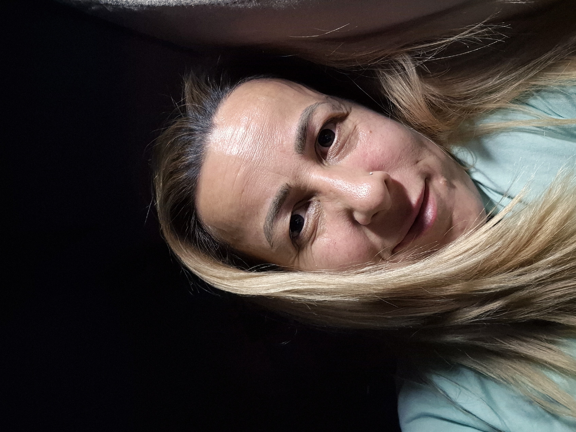 A close-up, selfie-style photograph features a woman with blonde hair against a dark background. The woman's face is illuminated from the front, with shadows present on the left side of her face and the background. Her eyes are dark, and she has a slight smile with her lips parted, revealing her teeth. She is wearing a light teal-colored shirt. A small nose ring is visible. The overall lighting creates a mood that is neither overtly bright nor completely dark, with a focus on her facial features. The location is not specified.
