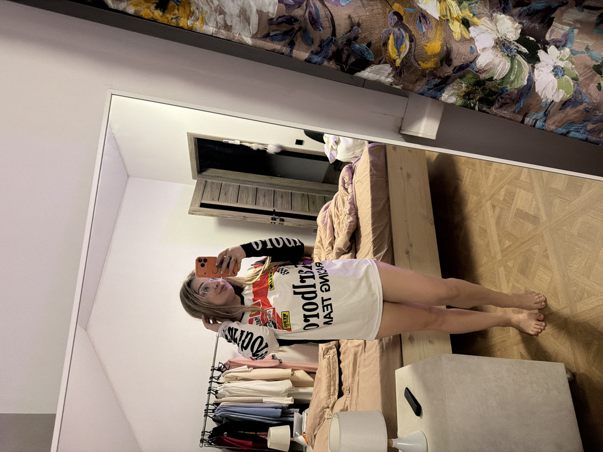 A young woman is captured in a full-body mirror selfie taken in what appears to be a bedroom. The original image is rotated 90 degrees clockwise; when viewed upright, she stands facing the mirror.
The woman has light brown hair and wears clear-framed glasses. She holds an orange smartphone in her right hand, positioned to take the photo, and looks directly at the phone's camera. She is wearing a long-sleeved white top or t-shirt dress with racing-themed graphics. The text on her shirt, visible in reverse in the mirror, clearly reads "MALBORO RACING TEAM" across the chest and "GOOD YEAR" below it. The sleeves also display "MALBORO" in white text against a black background. She is barefoot, and her toenails are painted.
The setting is an indoor room with light-colored walls and a light wooden floor featuring a herringbone or parquet pattern. To the woman's left, a clothing rack is visible, holding a variety of garments. Behind her, a bed with light, possibly pink or beige, bedding is partially visible. Above the bed, a patterned fabric, possibly a duvet or curtain, with floral and bird motifs in shades of brown, green, white, and yellow, hangs or is draped. A small, light-colored square stool or ottoman with a black handle is on the floor near her feet. In the reflection behind the woman, a wooden cabinet or drawer unit with slatted doors can be seen, along with what appears to be a roll of toilet paper and a lampshade. The lighting suggests it is daytime.