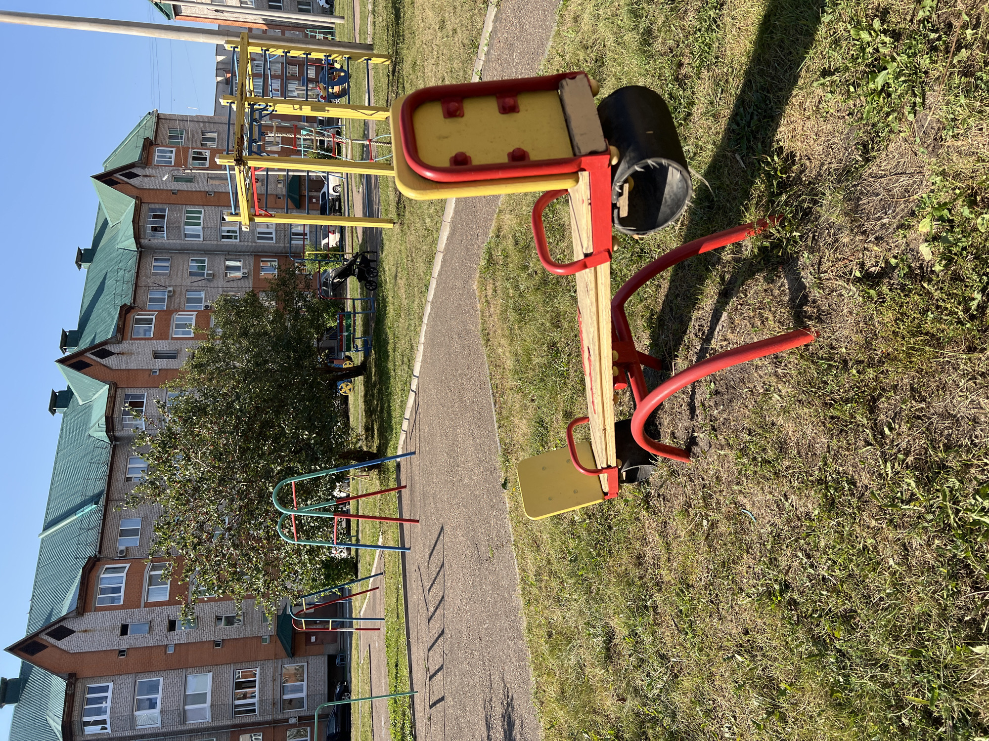 The image depicts a playground in the city of Salavat, Russia. In the foreground, a bright red and yellow seesaw with worn black cylindrical ends is positioned on a grassy area. Behind the seesaw, a paved pathway leads towards a complex of apartment buildings with distinctive green, pointed roofs and grey brick facades. Several other playground structures, including a swing set with red and blue supports and a yellow multi-level jungle gym, are visible in the midground. A tree with green foliage stands between the seesaw and the apartment buildings. The sky is clear and blue.