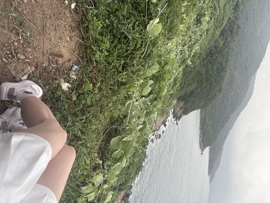 The image depicts a first-person perspective from a vantage point on a vegetated hillside overlooking the coastline of Sơn Trà Ward, Vietnam. In the foreground, the legs of an individual are visible, wearing light-colored shorts or a skirt, white socks, and white athletic shoes with light purple accents. The individual appears to be seated or resting on a dirt patch littered with small pieces of trash, including plastic wrappers and paper. Immediately beyond this patch, dense green foliage, including various grasses and leafy vines, covers the steep slope leading down to the sea. The coastline features a rocky shore where the ocean water, appearing grayish-blue and slightly choppy, meets the land. In the background, multiple layers of lush green, heavily forested mountains extend along the coast and recede into the distance under an overcast or cloudy sky.