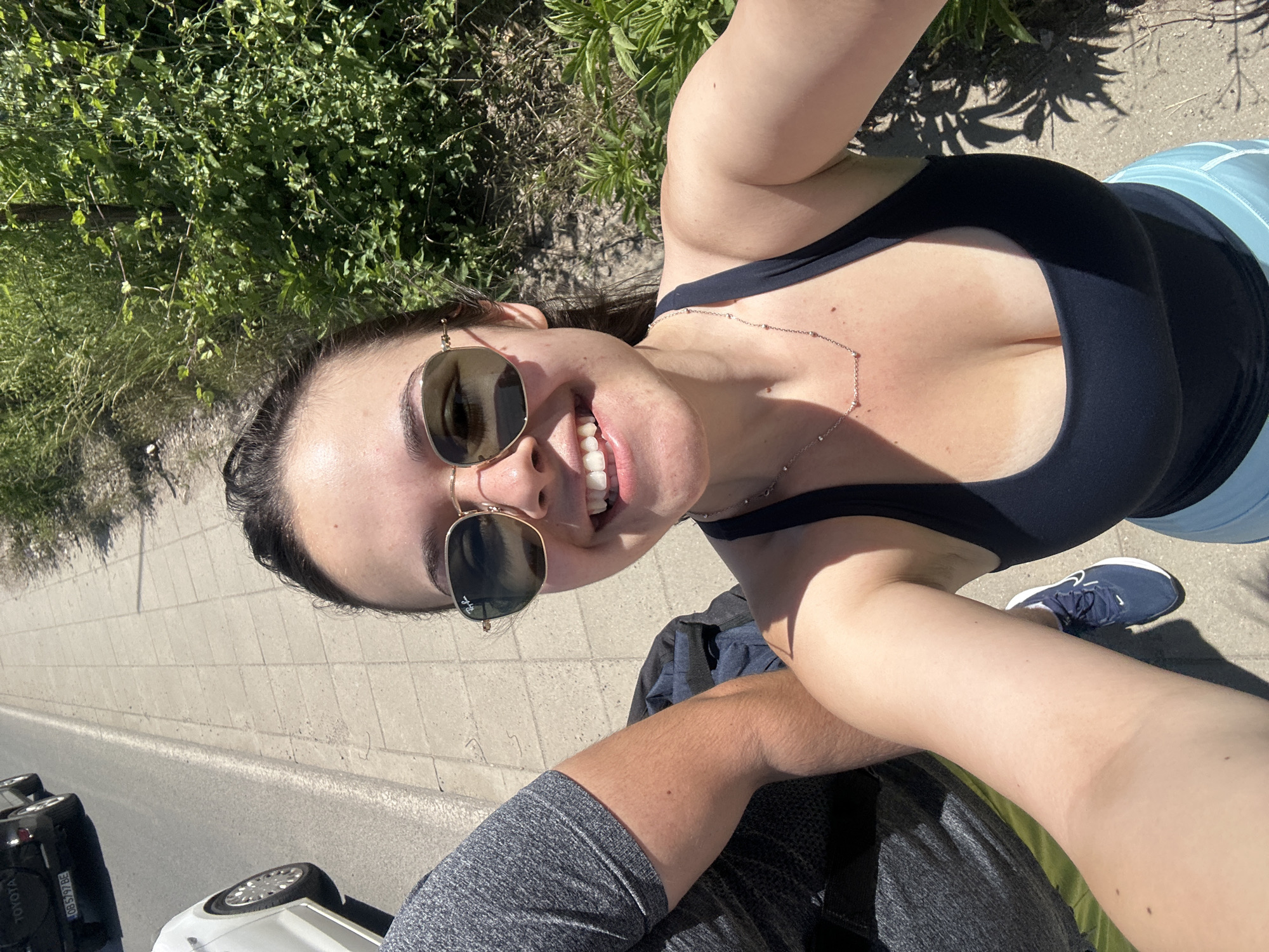A woman with fair skin and dark hair pulled back is wearing sunglasses and smiling broadly. She is wearing a dark sports bra and light blue shorts, along with a delicate silver necklace. She is positioned close to the camera, suggesting she's taking a selfie. The background shows a sidewalk or road, a patch of green vegetation, and a portion of a white car with the word "TOYOTA" visible on its license plate, potentially indicating the location as Sofia, Bulgaria. A man's arm and part of his gray shirt are also visible, suggesting he may be sitting next to her.