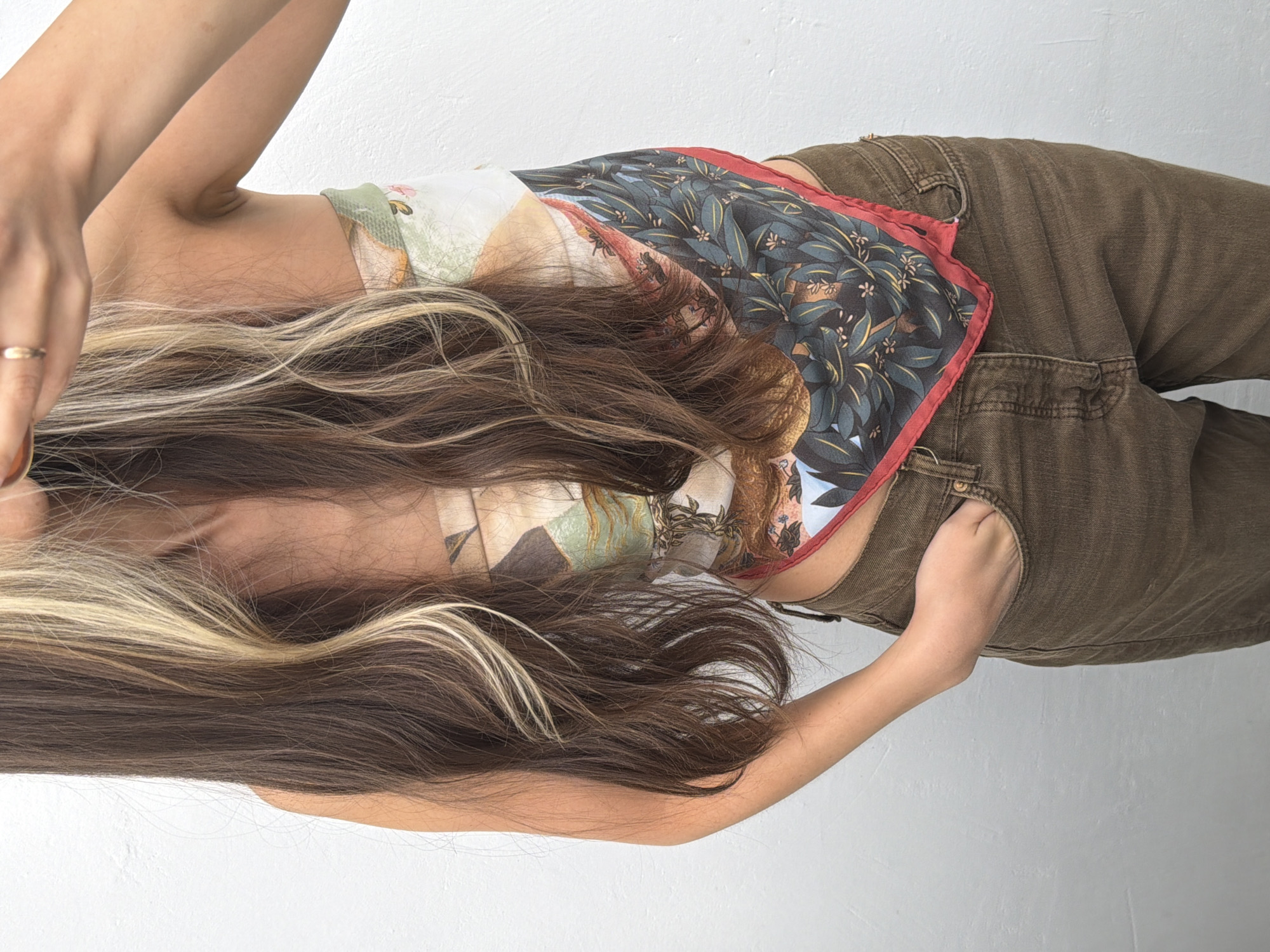 This is a top-down, close-up studio shot of a person lying on a white surface. The person has long, dark brown hair with blonde streaks, which cascades down their back and covers part of their torso. They are wearing a colorful, patterned top with a bandana-like design and olive-green pants. A gold ring is visible on their left hand. The lighting is bright and even, suggesting artificial light or a well-lit indoor space. The background is a plain white wall. The focus is on the texture and patterns of the clothing and hair, and the overall mood is neutral and aesthetic.