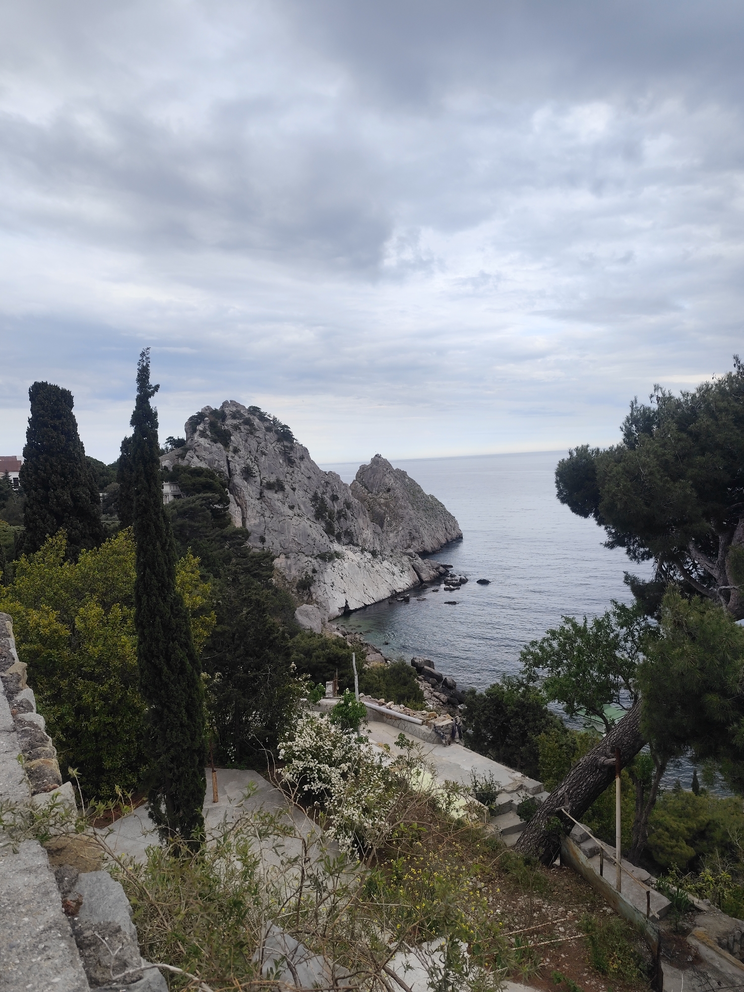 The image captures a coastal scene on an overcast day. A rocky promontory extends into the sea, with various trees and vegetation dotting the landscape. A built structure, possibly a pathway or terrace, is visible in the foreground, partially obscured by foliage. The scene is characterized by natural elements, including the sea, rocks, and plant life.