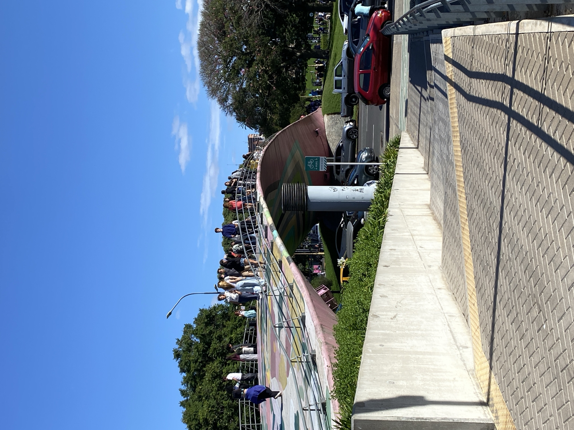 The image depicts the Curutchet Overpass, located in Buenos Aires, Argentina. The overpass, a pedestrian bridge decorated with colorful murals, is crowded with people standing along the railing. Cars are parked and driving alongside the bridge. Trees are visible behind the overpass and in the background. The sky is blue with few clouds. The lower right side of the image shows a sidewalk with textured pavement and a concrete barrier. Shadows from the overpass railing are cast on the sidewalk. A lamppost is on the left side of the image.