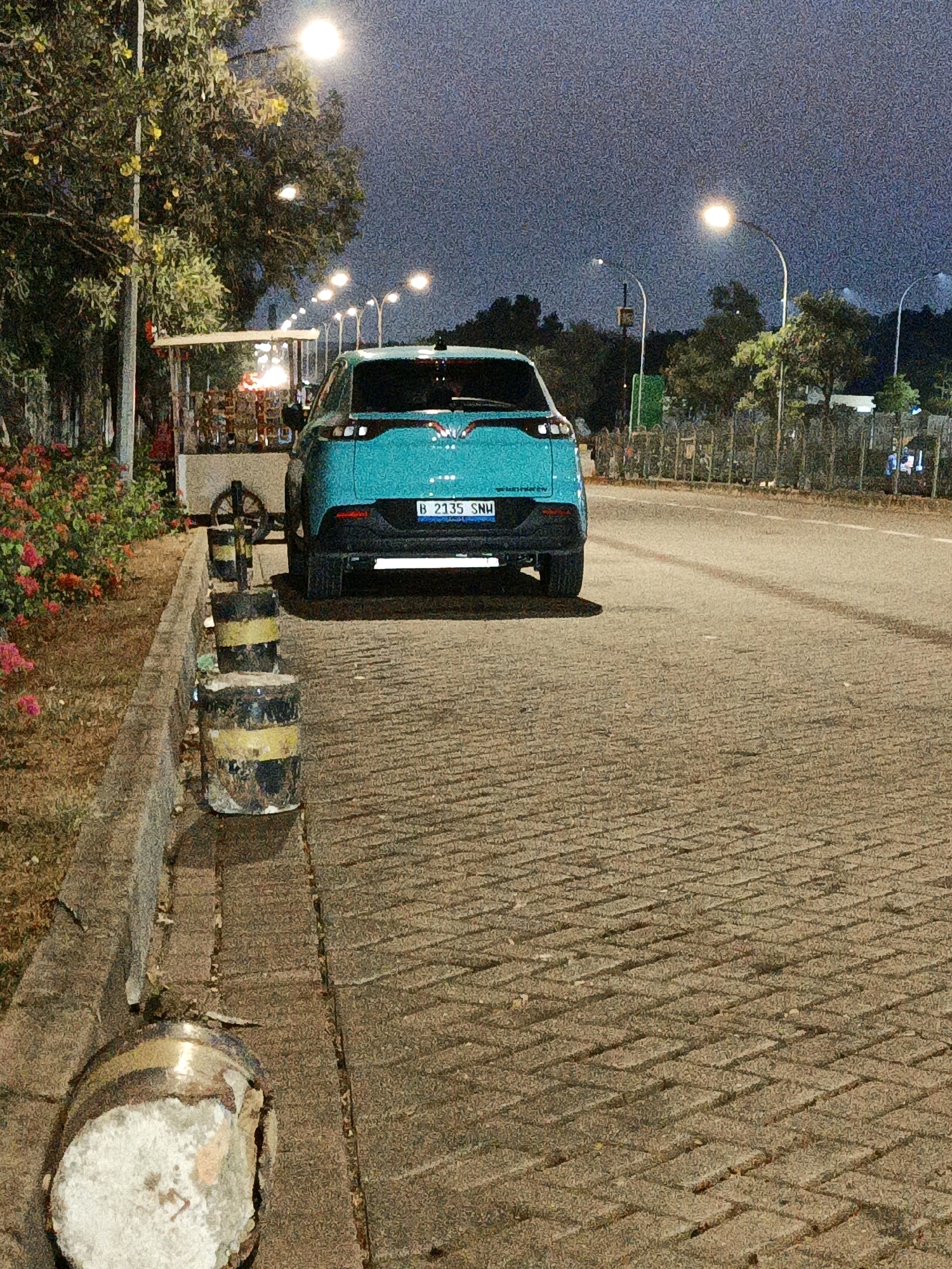 The image captures a street scene at dusk in North Jakarta, Indonesia. In the foreground, a teal-colored car with license plate B 2135 SNU is parked on a paved road. To the left of the car, a sidewalk is lined with concrete barriers painted with yellow and black stripes, and further left, a small cart is visible, possibly selling food or goods. The environment includes trees, flowering plants, and streetlights illuminating the area. The sky is a dark, dusky blue.