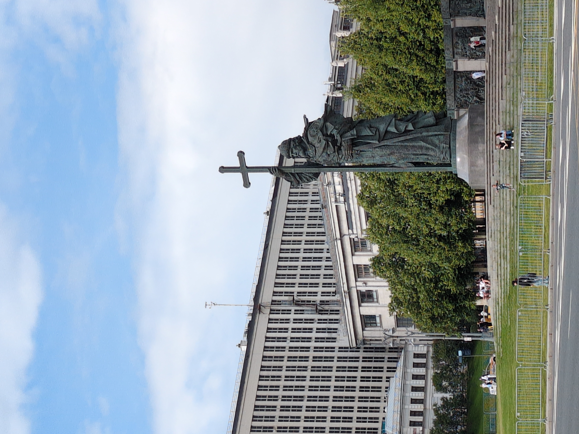 An outdoor, eye-level, wide shot captures the bronze statue of Christopher Columbus on a pedestal in the city of Unknown, Unknown. The statue is positioned on the right side of the image, dominating the center of the frame. Columbus is depicted in full figure, standing upright and holding a large cross in his right hand, extending it upwards. He is dressed in historical attire, including a helmet, armor, and a long cloak that drapes down his body. The statue is a dark bronze color and appears weathered.
Behind the statue, a large, contemporary building stretches across the background, characterized by a grid of numerous windows. To the left of the statue and in front of the large building, a smaller, older-style building with visible windows and architectural details can be seen. Lush green trees form a dense line between the buildings and the statue, creating a natural backdrop.
In the foreground, a grassy area is visible, separated from a road by a metal fence. Several people are scattered throughout the scene. On the grass in front of the statue, a few individuals are sitting on steps leading up to a lower level of the monument. Further in the foreground, near the fence, a single person stands, looking towards the statue.
The sky above is a pale blue with scattered white clouds, indicating fair weather. The overall scene suggests a public space, possibly a park or plaza, where the monument is a central attraction.