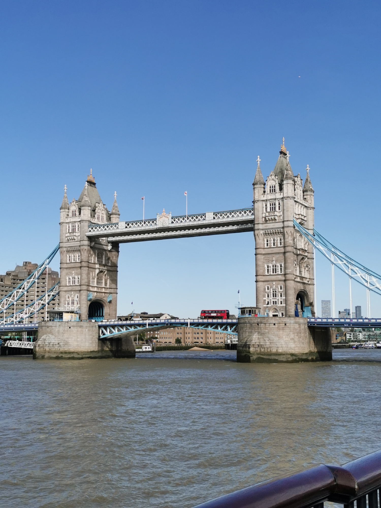 The image presents a wide, clear view of the iconic Tower Bridge spanning the River Thames in London, United Kingdom, on a bright, sunny day. The bridge, constructed from light grey stone with intricate Gothic Revival architectural elements, features two prominent towers connected by high-level walkways and a lower roadway. White and blue suspension cables are visible on both sides of the bridge, anchoring it to the banks.
A classic red double-decker bus is captured crossing the bridge from right to left, adding a characteristic London element to the scene. Buildings line both sides of the river in the background, with modern structures visible on the left and more traditional brick buildings on the right. The River Thames occupies the lower half of the frame, its surface showing gentle ripples and appearing a brownish-grey color. A dark, ornate railing is partially visible in the bottom right corner, indicating the vantage point of the photographer.
The sky is a brilliant, cloudless blue, suggesting fair weather and good visibility. A tiny, faint white speck can be observed high in the sky, possibly a distant aircraft. The scene captures a typical daytime moment at a famous landmark, devoid of any specific event beyond the general flow of traffic and river activity. No significant text is clearly legible, though the bus has a small "15" on its front.