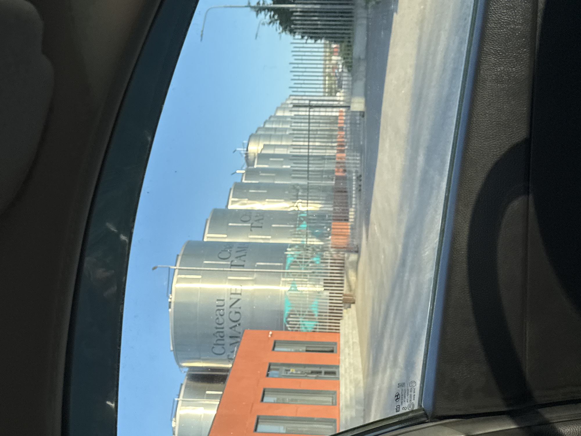 The image shows a row of large, cylindrical, silver storage tanks behind a fence, reflecting the sky and buildings, as seen through a car window. The tanks have writing on them, including "Château TAMAGNE". A red building is visible in front of the tanks. The location is likely Taman', Russia, based on the brand name. The foreground shows the interior of a vehicle, with the window frame and dashboard visible. The overall impression is of an industrial area viewed from inside a car. There are no people visible.