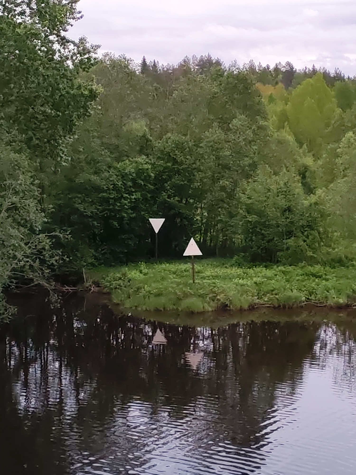 The image depicts a water body (likely a river or lake) with dense green vegetation on the opposite bank. Two triangular white signs are visible on the bank, one pointing up and the other pointing down. The signs are positioned on posts. The water surface reflects the trees and signs, creating a mirrored effect. The background shows a forest. The location is potentially Volokov Most, Russia, based on the prompt.