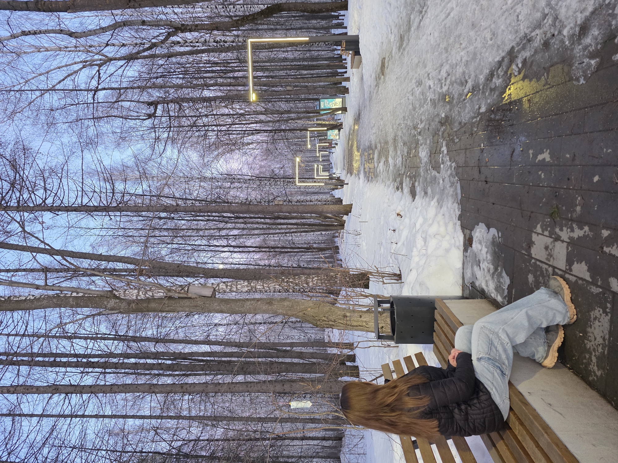 A girl sits on a park bench. The location is Chaikovsky, Russia. The park is lined with bare trees and rectangular light posts on the right side. There is snow on the ground. The path is brick. The girl has long hair, is wearing a black puffer jacket, blue jeans, and tan boots. A black trash can is to the left of the bench.