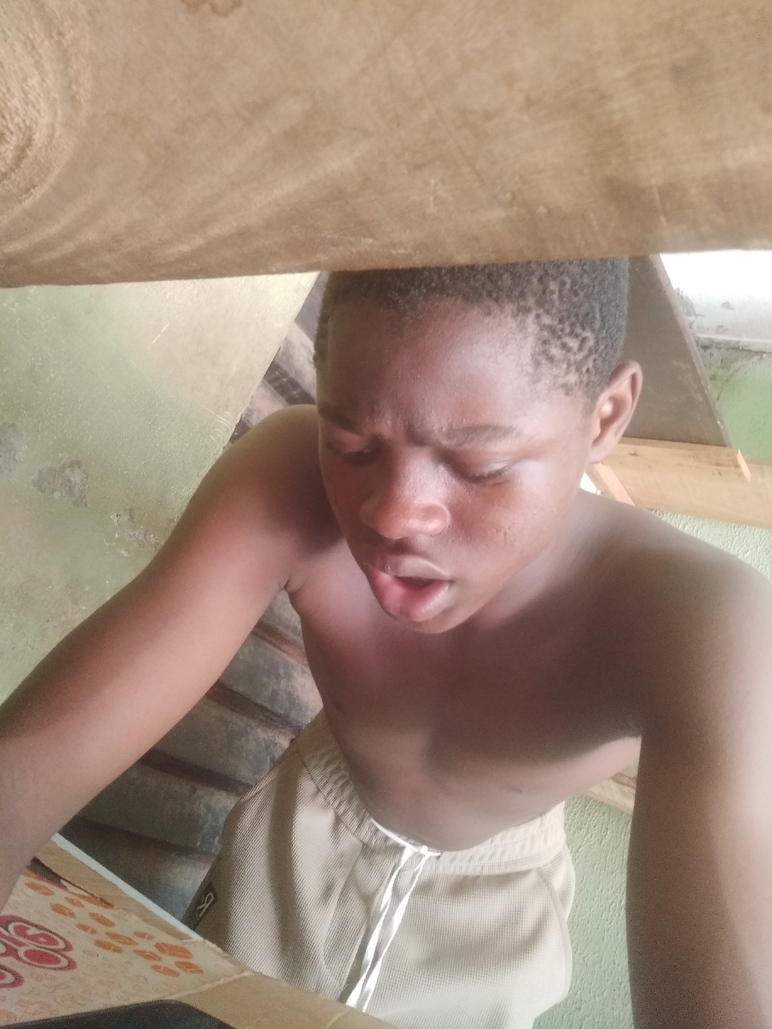 A young person, likely male, with dark skin and short, dark hair is captured in a close-up, slightly upward-angled shot. He is shirtless and wears light-colored shorts with a white drawstring. His head is tilted slightly down and to the left, and his eyes are mostly closed or looking downwards intently. His mouth is open, as if in mid-speech, gasping, or concentrating.
A large, light-colored wooden plank or beam fills the top quarter of the frame, partially obscuring the view from above. Below the subject, on the left, a surface with an orange and red abstract pattern is visible, possibly a mat or tablecloth. The background features a light green or pale yellow wall, which appears somewhat textured. In the upper right corner of the background, additional wooden structures, possibly part of a roof or support beams, are visible. The lighting suggests it is daytime. There is no discernible text visible in the image. The overall setting appears to be a simple indoor or semi-enclosed space.