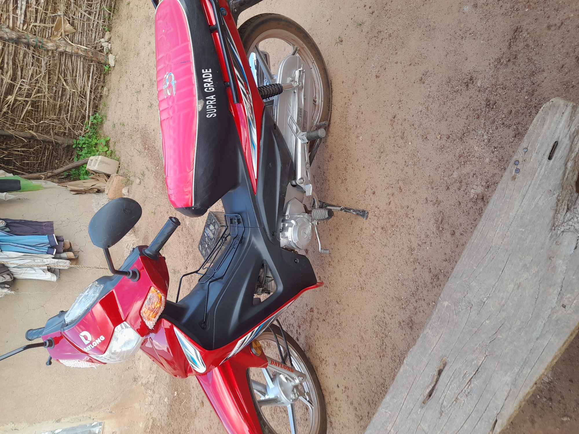 A red and black motorcycle is parked on a dirt ground in Ibi, Nigeria. The motorcycle has a red body with black accents and a red seat. A logo with the word "DAYLONG" is visible on the front of the motorcycle. The word "SUPRA GRADE" is printed on the side of the seat. The background shows a building with a thatched roof and some laundry hanging on a line. A wooden beam is in the foreground, partially obscuring the motorcycle. The overall scene appears to be an outdoor setting, possibly a residential area.