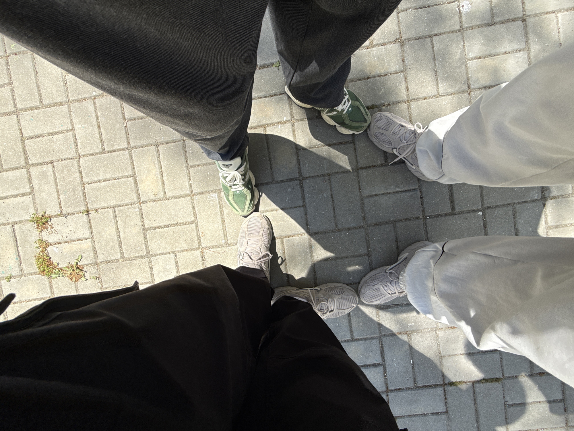 A high-angle, top-down photograph captures the feet and lower legs of four individuals standing on a grey paved surface, likely outdoors in Varna, Bulgaria. The subjects are arranged in a loose circle, facing inwards. One person wears dark grey jeans or trousers paired with green and white athletic shoes, featuring a visible 'N' logo. Another individual wears black trousers and light grey sneakers. The remaining two people are dressed in light grey or white trousers, both wearing similar light grey athletic shoes.
The ground is composed of rectangular grey pavers, with small patches of green grass or weeds growing in some of the cracks, indicating an outdoor, possibly urban or suburban, setting. Strong, well-defined shadows cast by the individuals across the pavement suggest bright sunlight, indicating a sunny day, likely during late morning or early afternoon. The activity appears to be a casual standing gathering, possibly paused for this specific perspective shot focusing on their footwear. No other text or specific events are discernible.