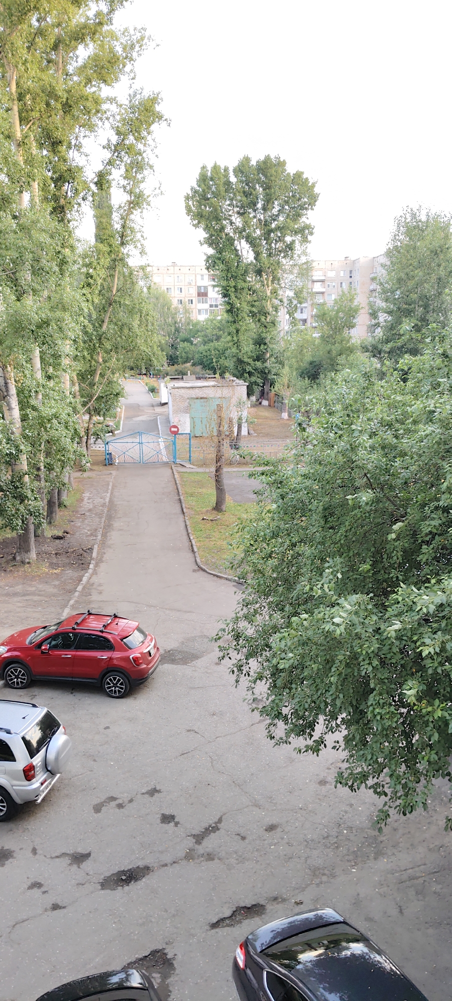 The image captures an aerial view of a street in the city of Zarinsk, Russia. A red SUV with a roof rack is parked on the asphalt road, next to a silver SUV. Further down the road, a blue gate with a red and white circular sign is visible, leading to a small brick building with a garage door. Trees line the sides of the road, and apartment buildings can be seen in the background. The sky is overcast and the lighting suggests it is daytime.